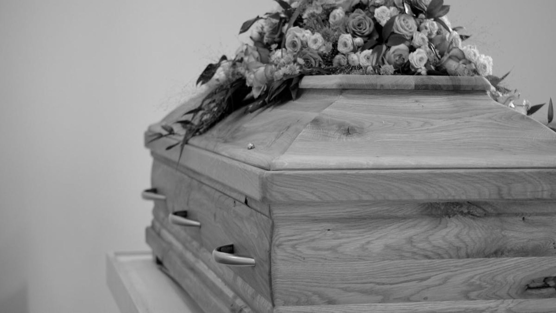 Un ataúd de madera coronado con un arreglo floral descansa sobre un pedestal en un entorno neutro.
