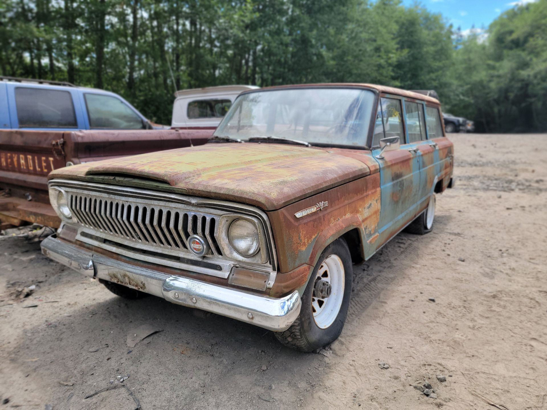 Rusty Jeep Wagoneer in a junkyard, showing heavy weathering on the body with trees in the background.