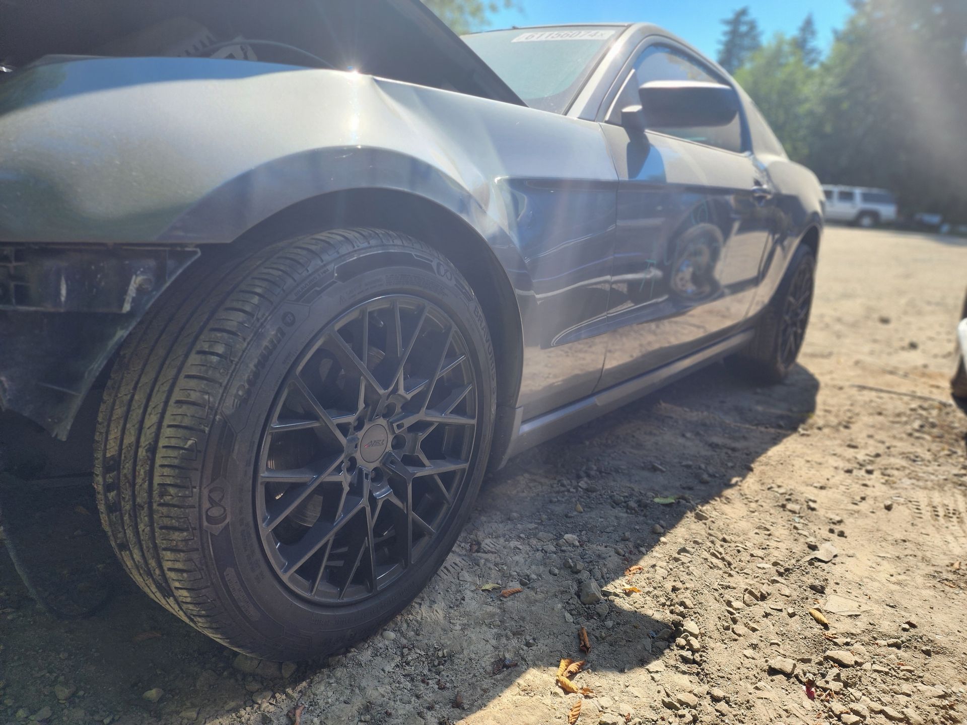 Dark gray Ford Mustang with damaged front, black rims, parked on gravel.