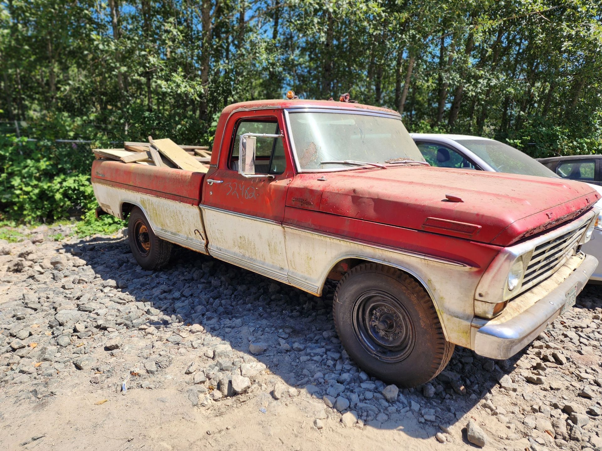 Red and white Ford pickup truck in disrepair, parked on gravel, near a forest.