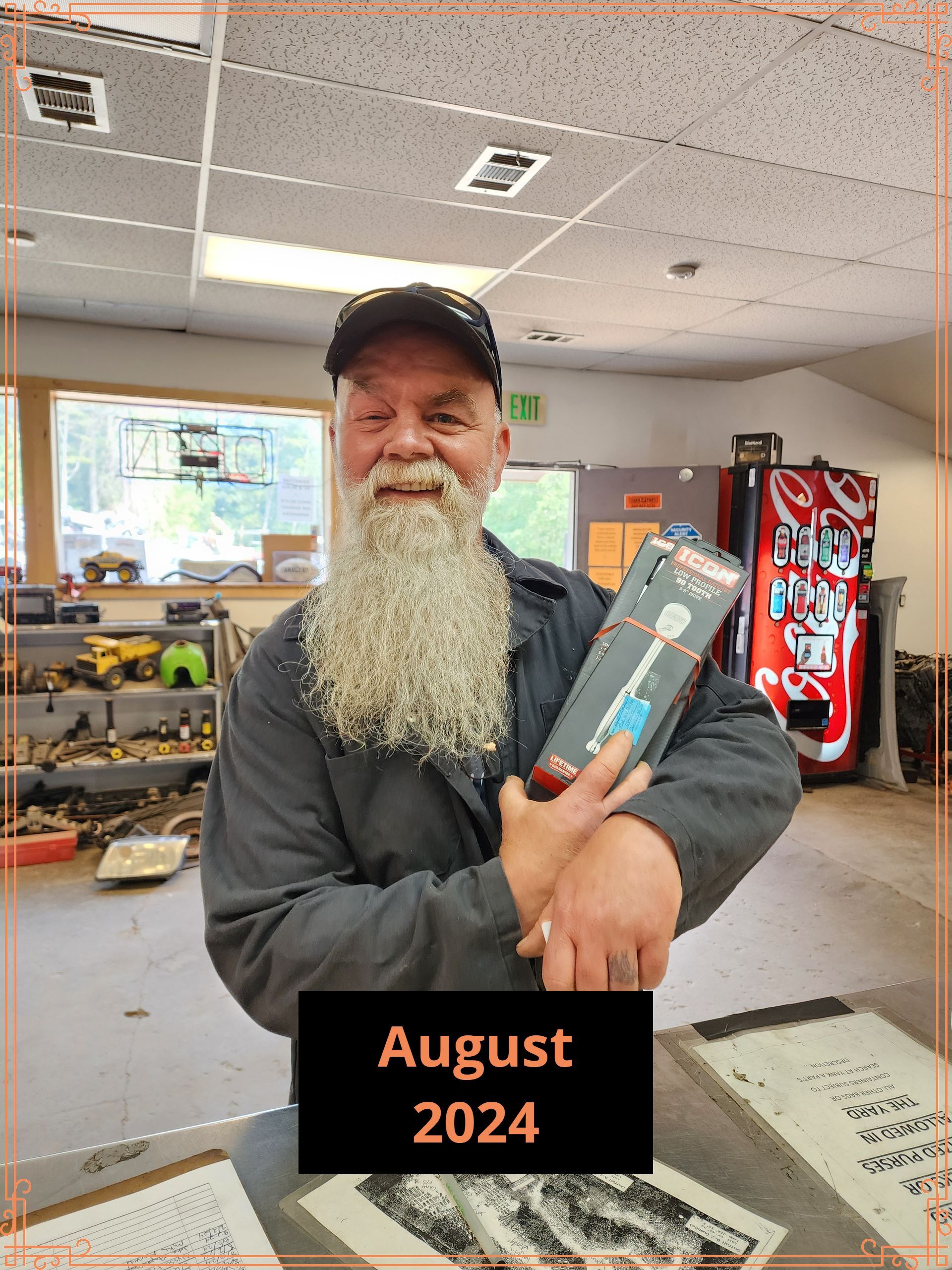 Man with long beard holds a box in shop. Sign in lower center says 