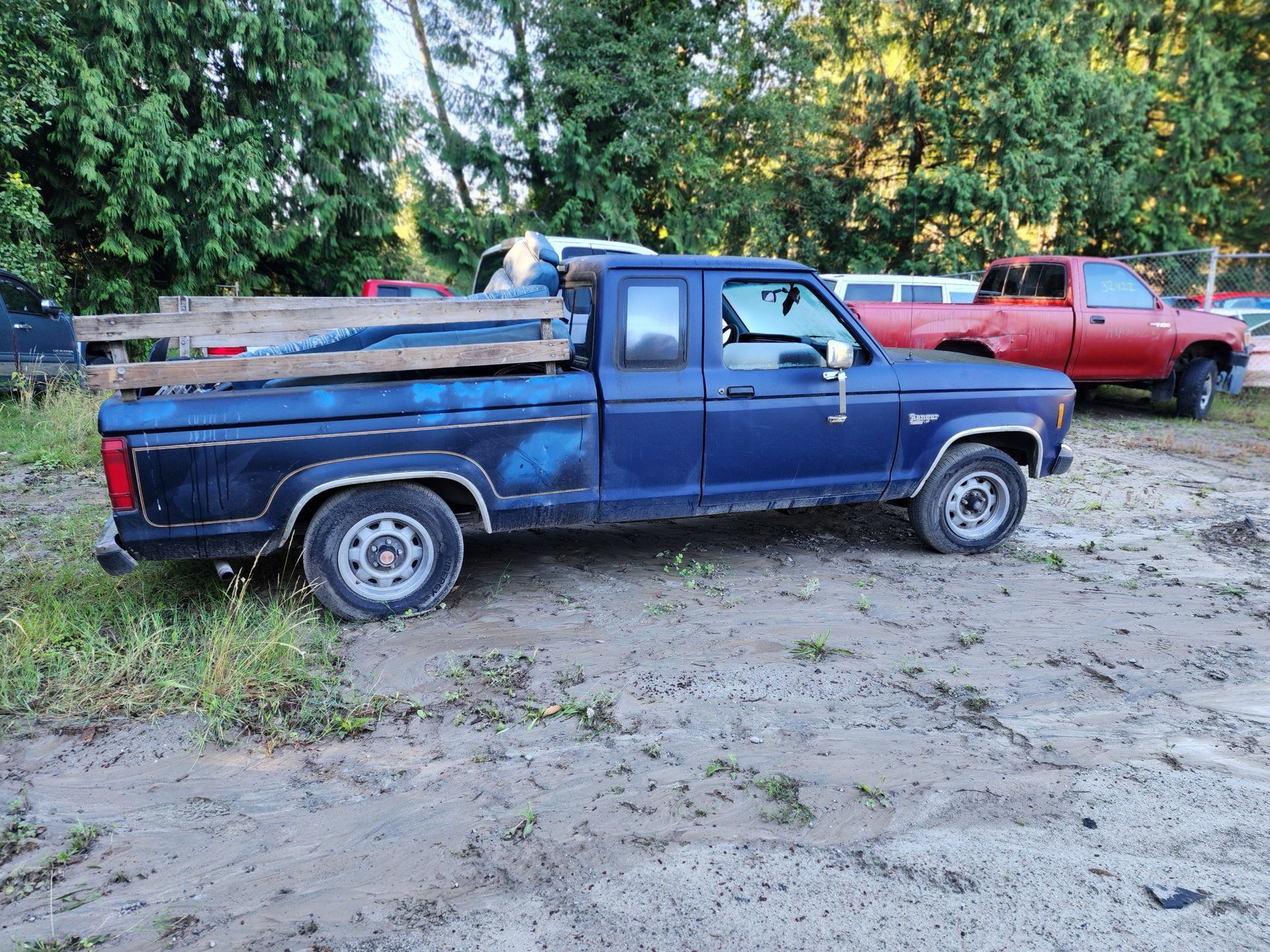 Blue Ford Ranger pickup truck with wooden planks in bed on a muddy lot, red truck in the background.