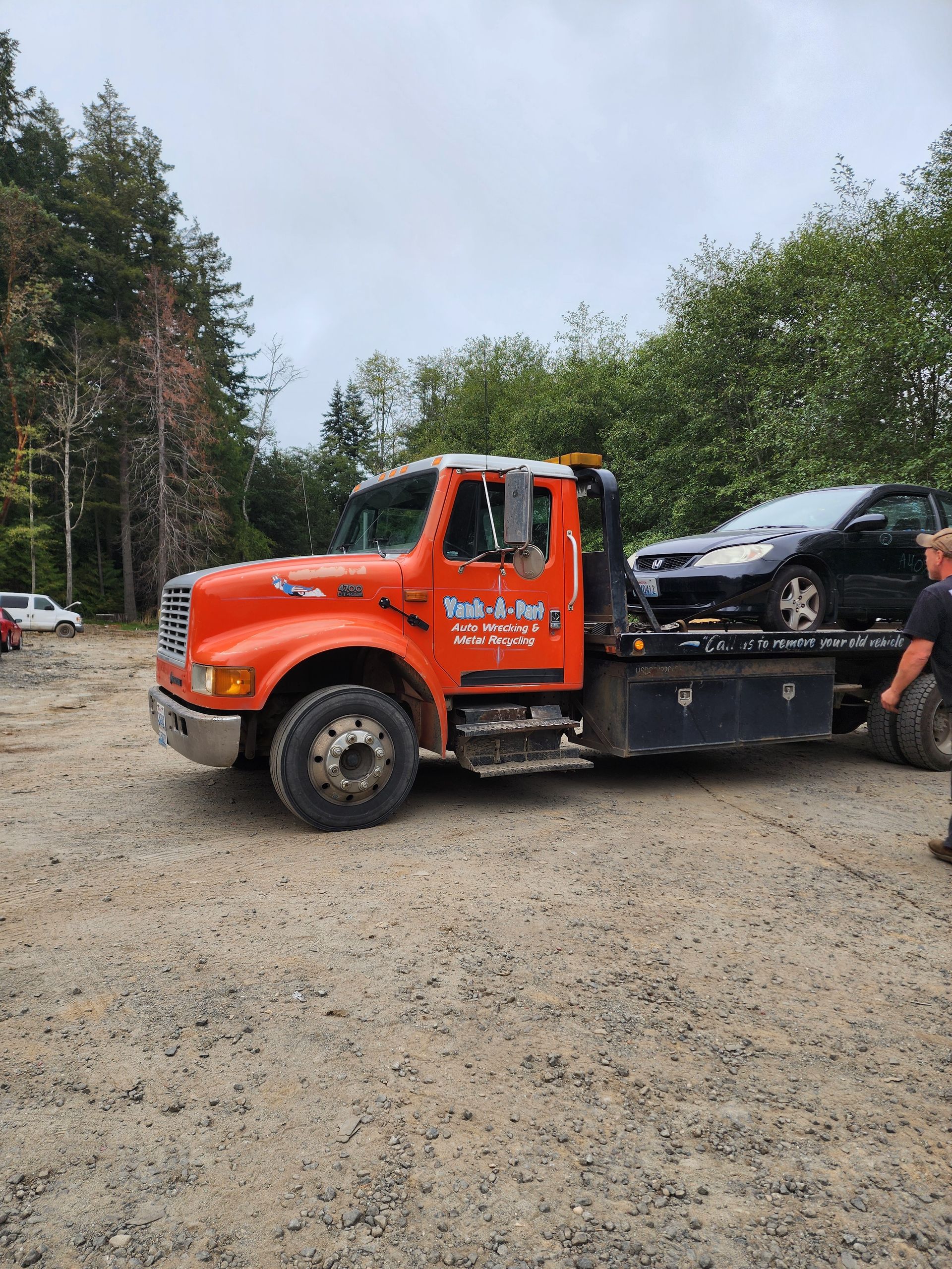 Orange tow truck towing a black car on a gravel lot with a cloudy sky and trees in the background.