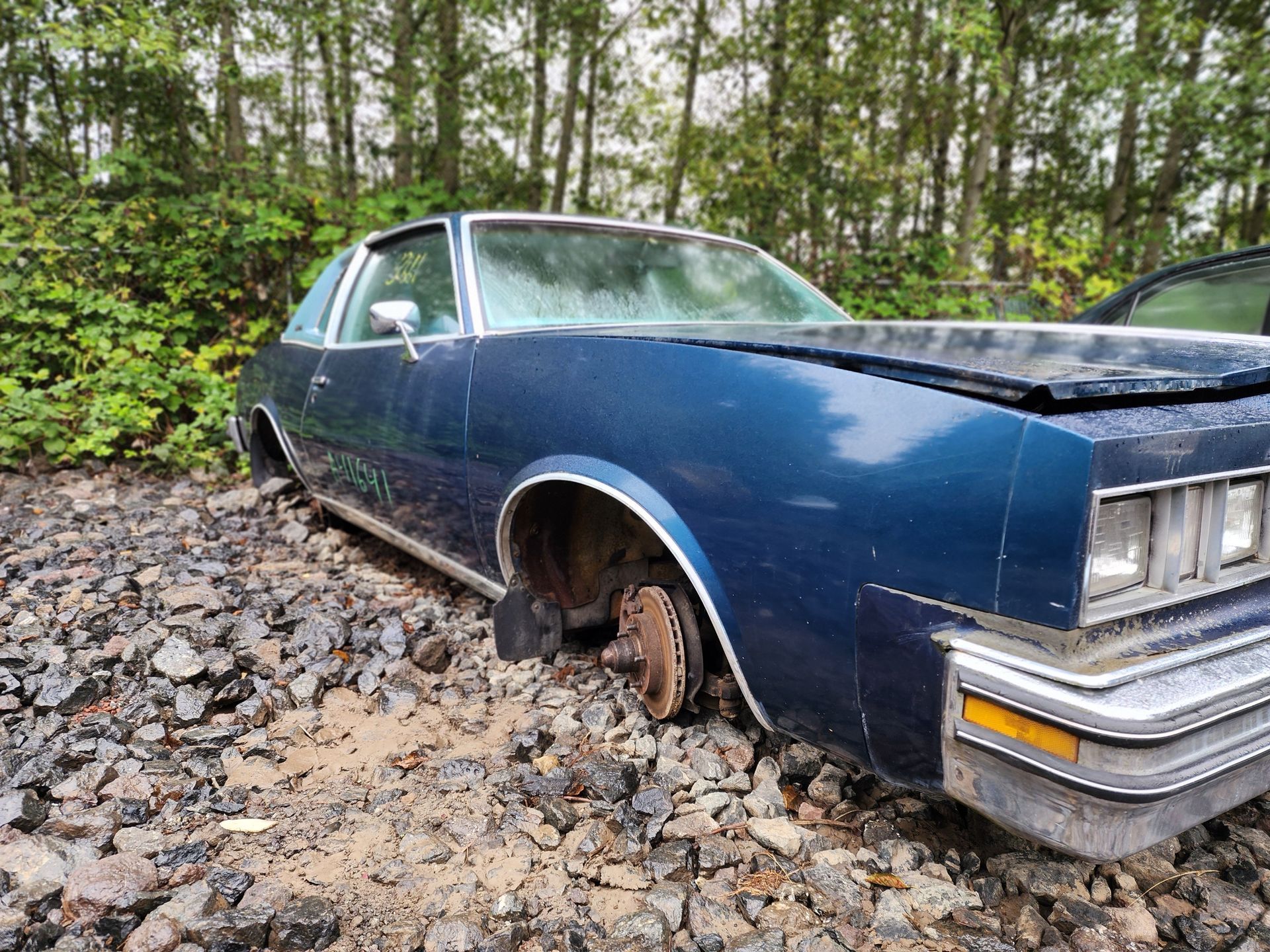 Blue, abandoned classic car with missing wheel in a wooded area.