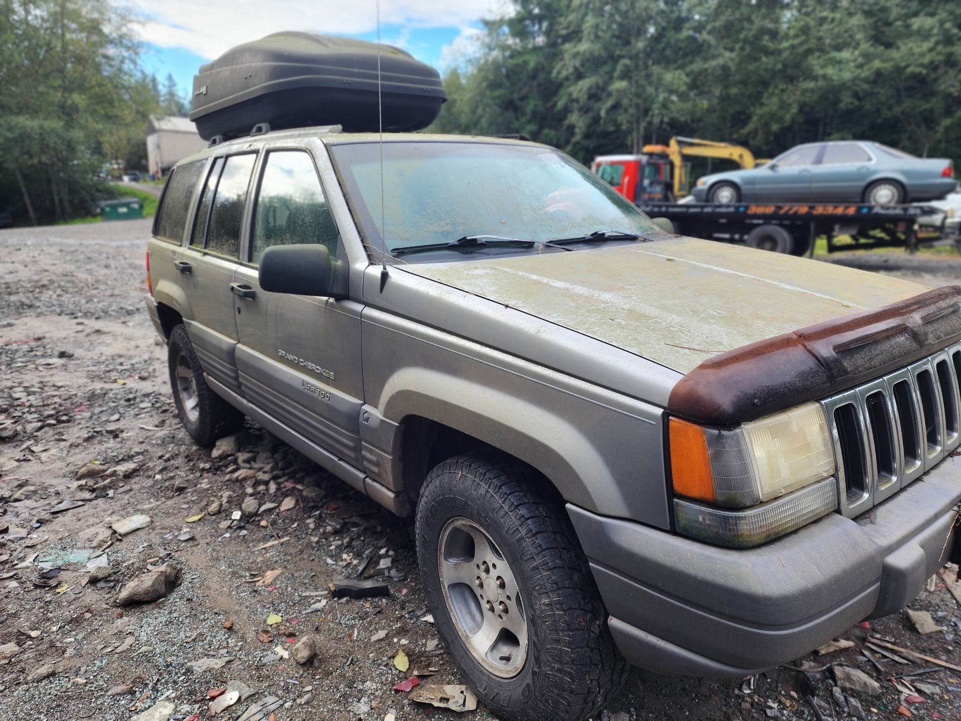 Gray Jeep Grand Cherokee with a cargo box on top, parked on gravel with a car on a tow truck in the background.