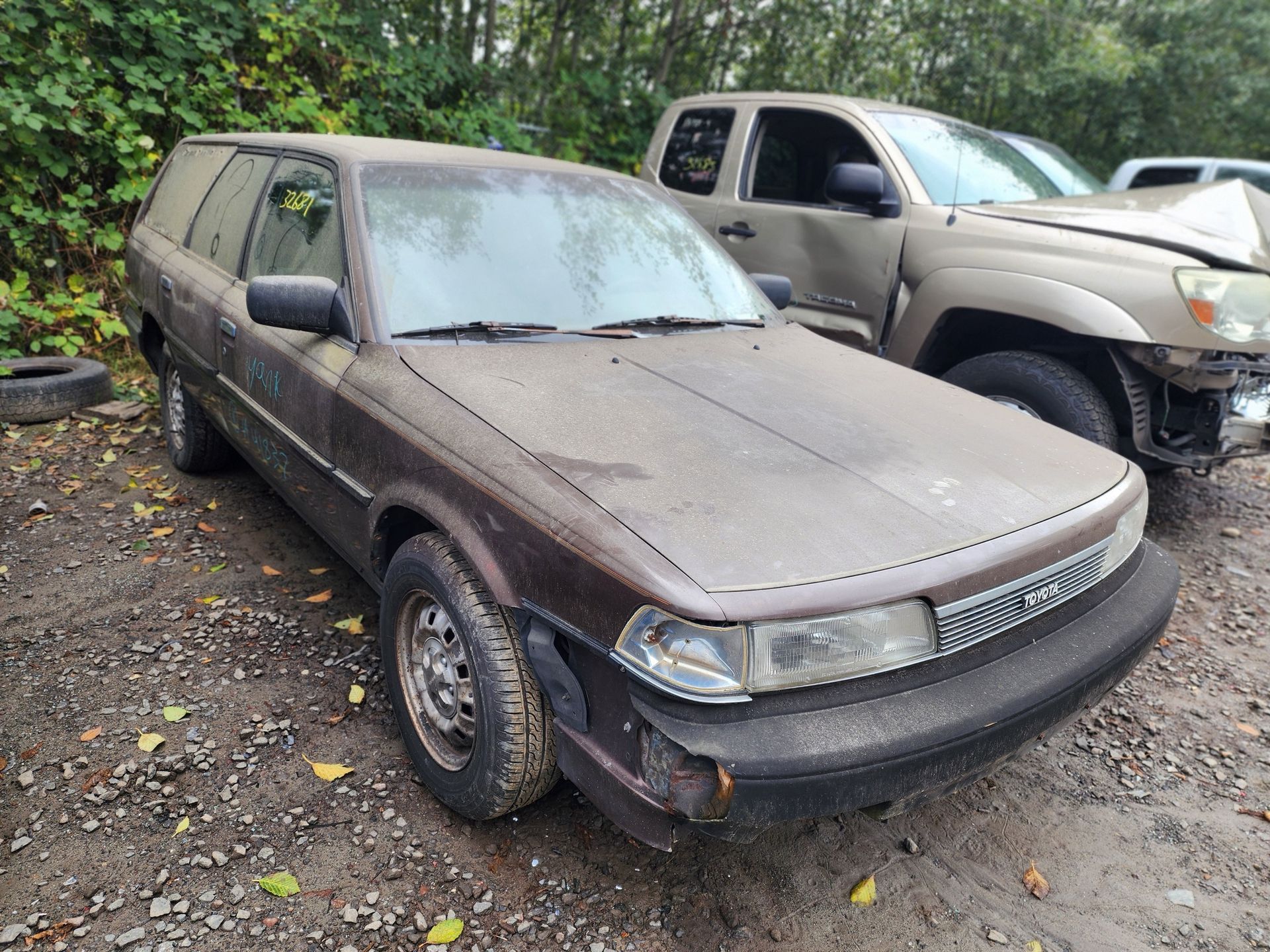 Brown Toyota Corolla wagon in a junkyard, covered in dirt, next to a damaged truck.
