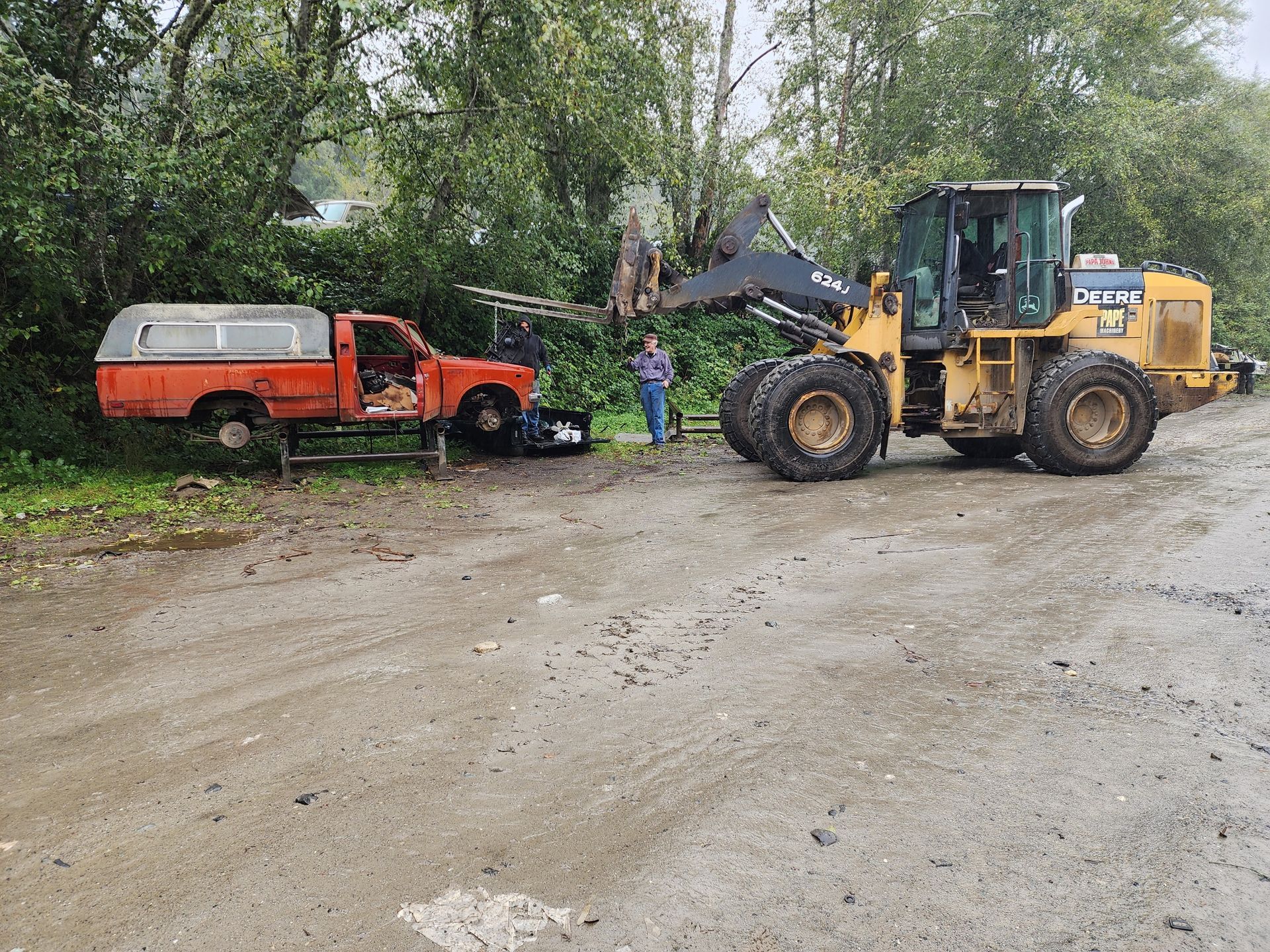 A yellow front-end loader pulls a derelict red pickup truck along a dirt road. A person stands nearby.