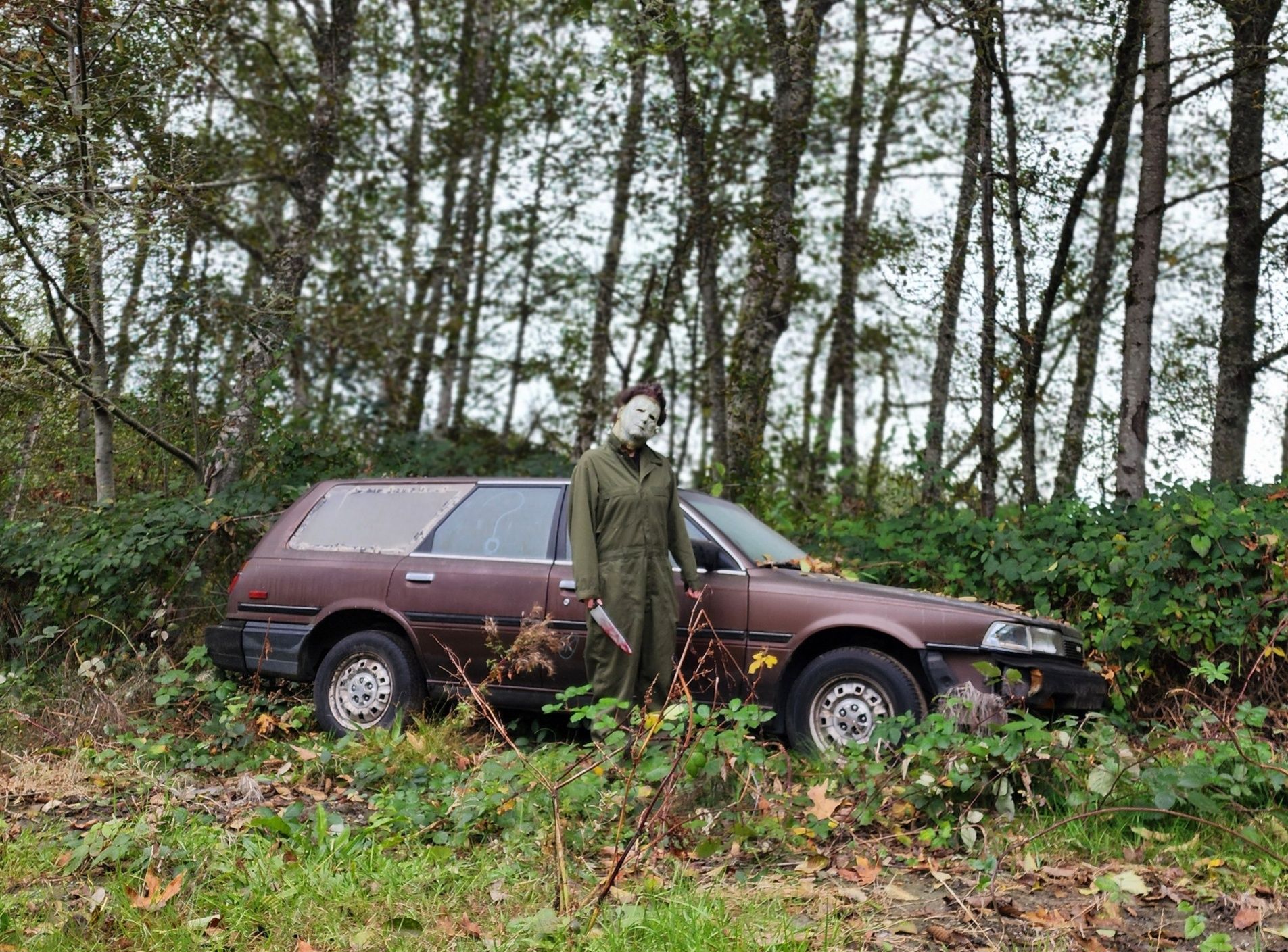 Michael Myers standing next to a brown station wagon in a wooded area.