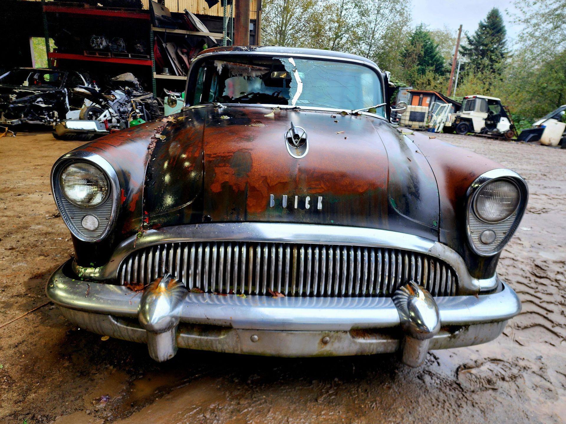 Rusty, vintage Buick in a salvage yard. Brown and black exterior, chrome accents, broken windshield.