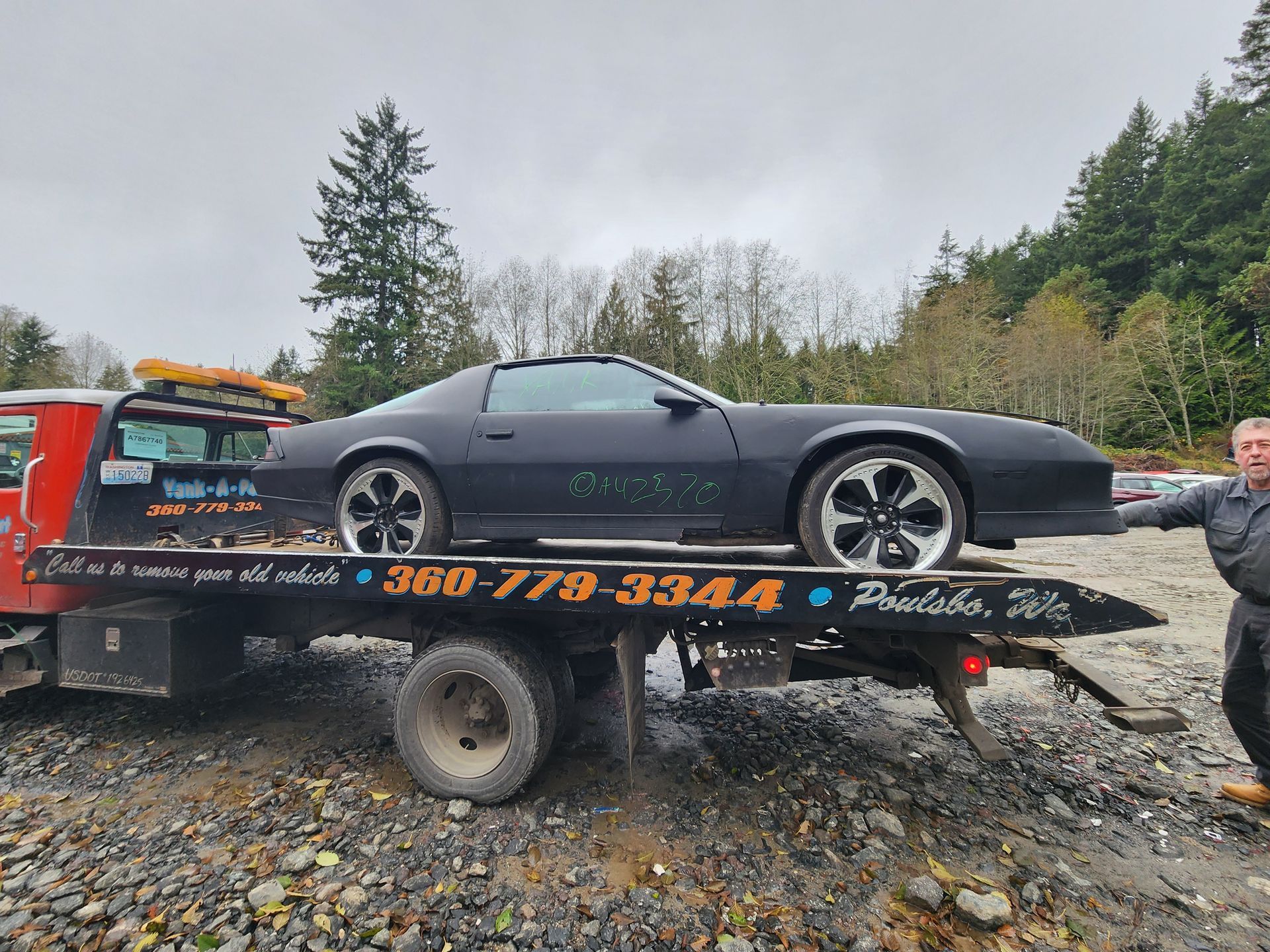 Black Camaro on a tow truck, man standing beside it. Cloudy day, outdoor setting.