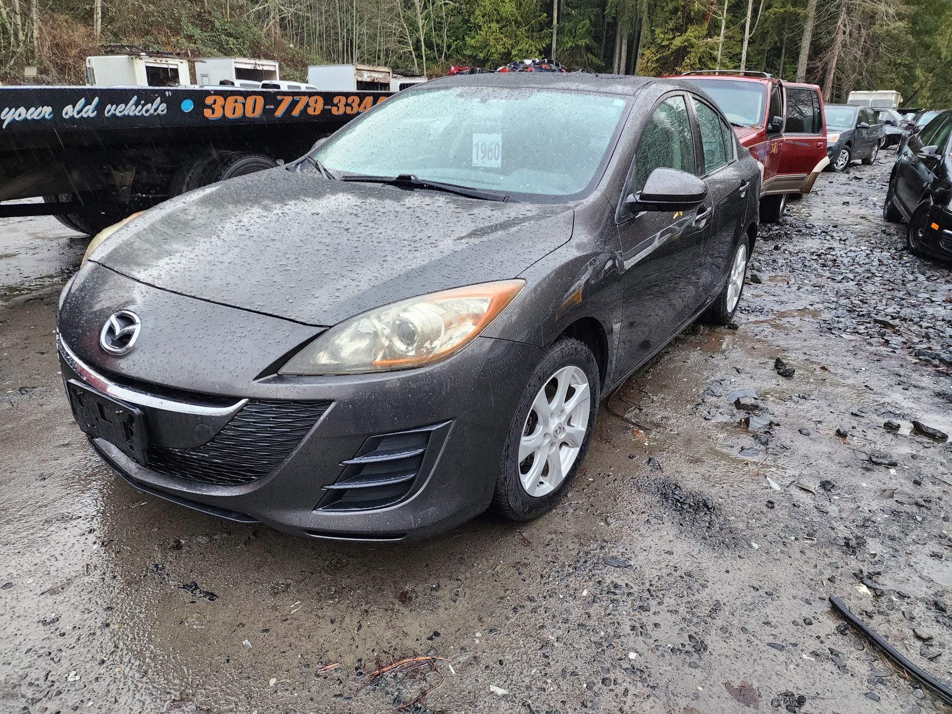 Gray Mazda 3 sedan in a muddy junkyard with other vehicles; cloudy day.