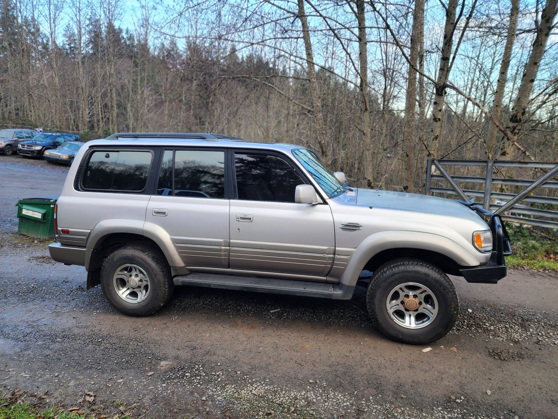 Silver Toyota Land Cruiser parked on gravel road, trees in background.