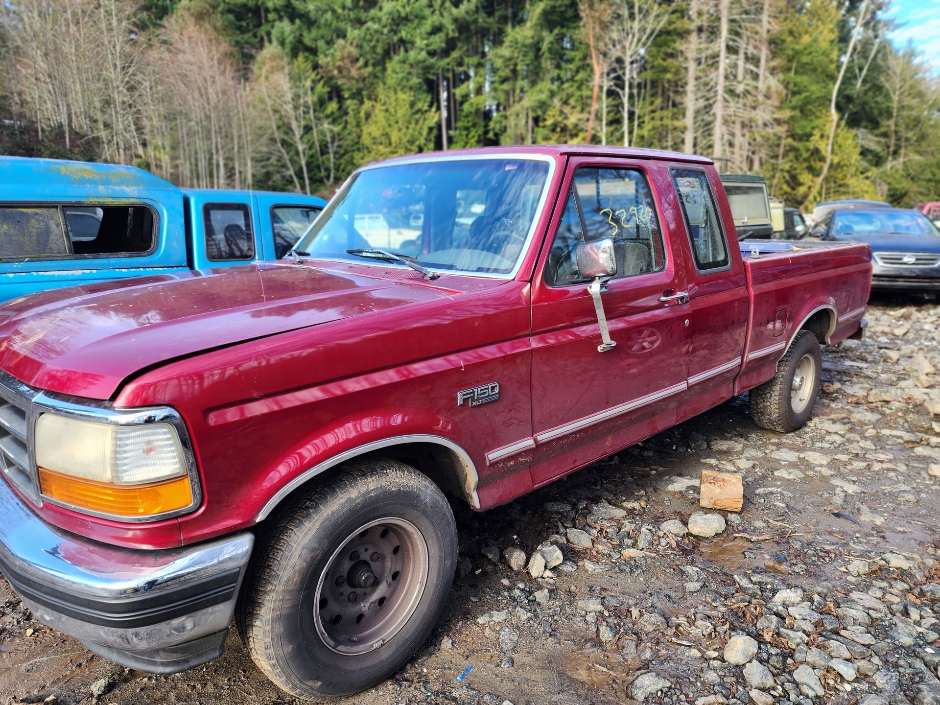 Red Ford F-150 pickup truck parked in a junkyard, surrounded by other vehicles and trees.