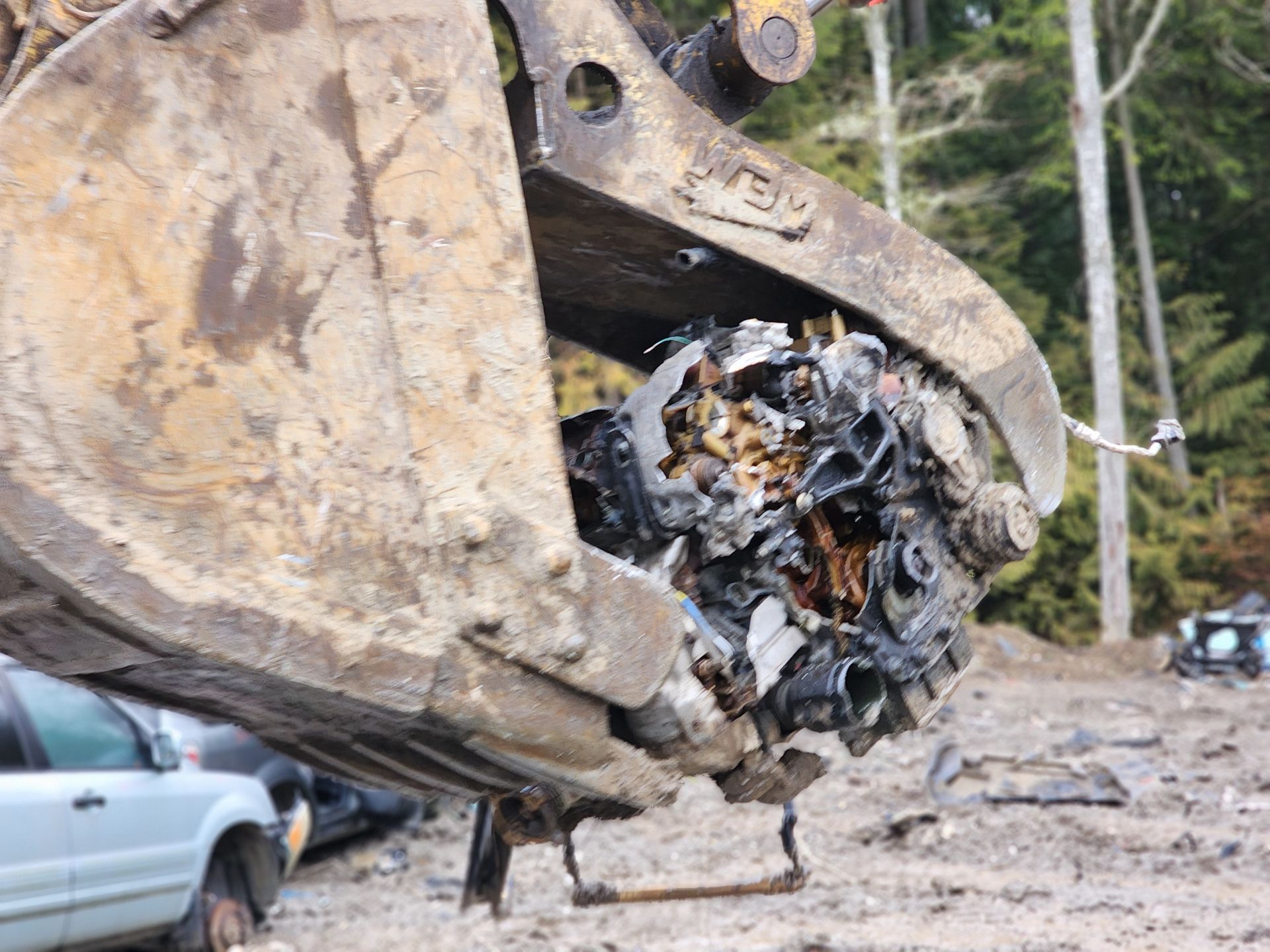 A large excavator bucket holding a crushed, burnt engine block at a salvage yard.