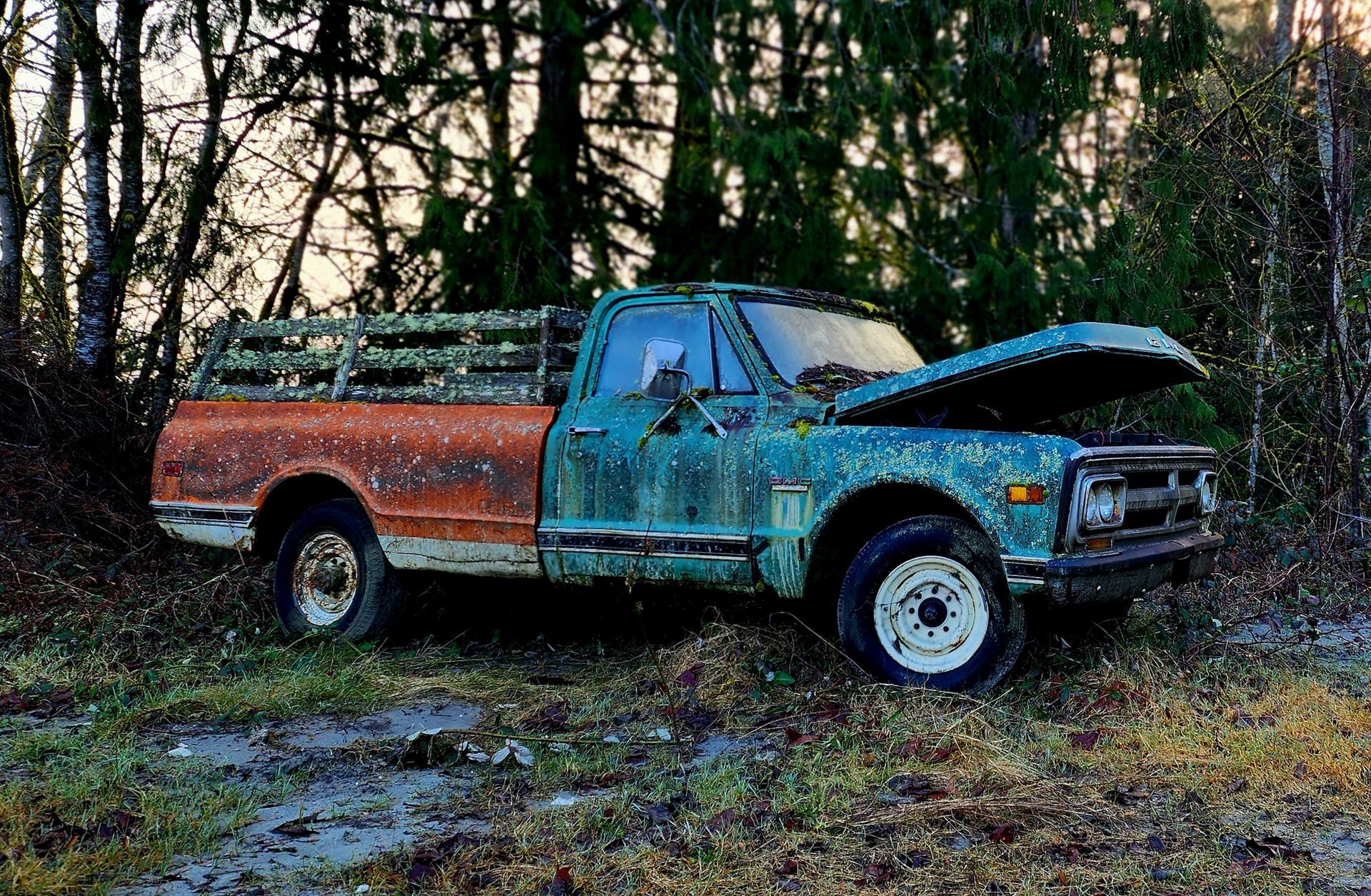 A weathered teal and orange pickup truck with the hood open, parked in a wooded area.