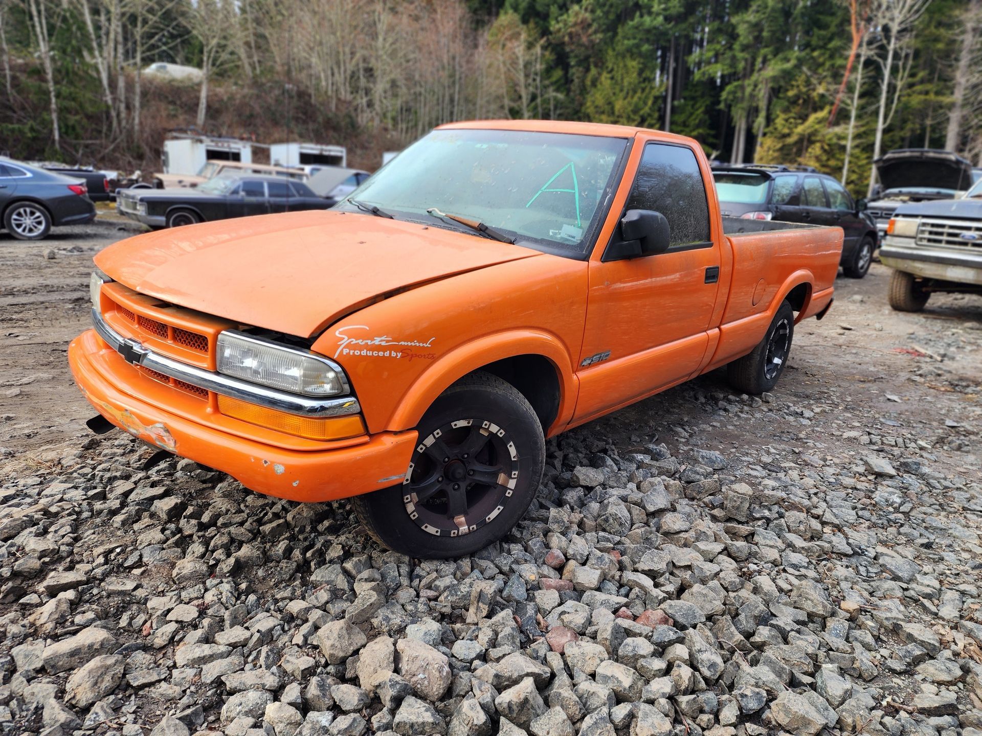 Orange Chevy S10 pickup truck parked in a rocky lot. The truck has black wheels and is in a salvage yard.