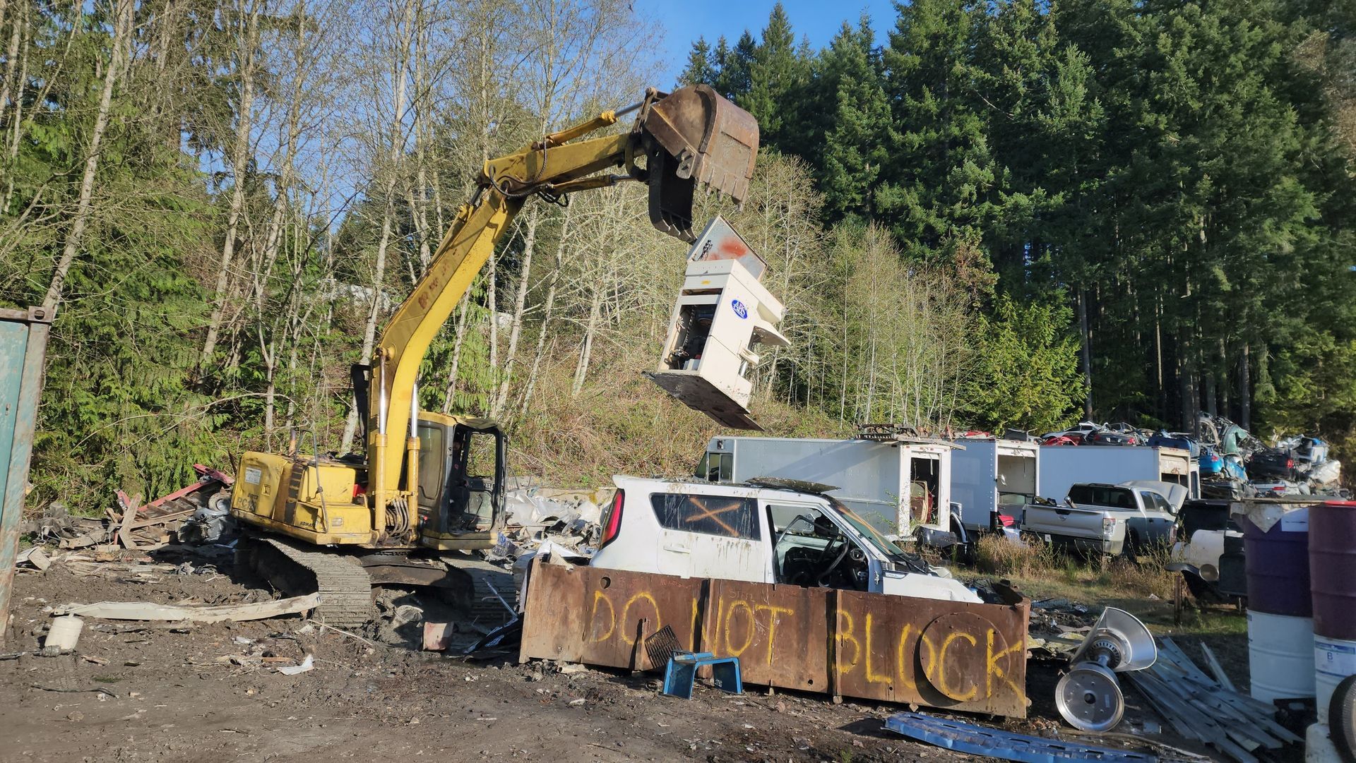 An excavator lifts a demolished structure at a salvage yard, with debris, vehicles, and trees in the background.