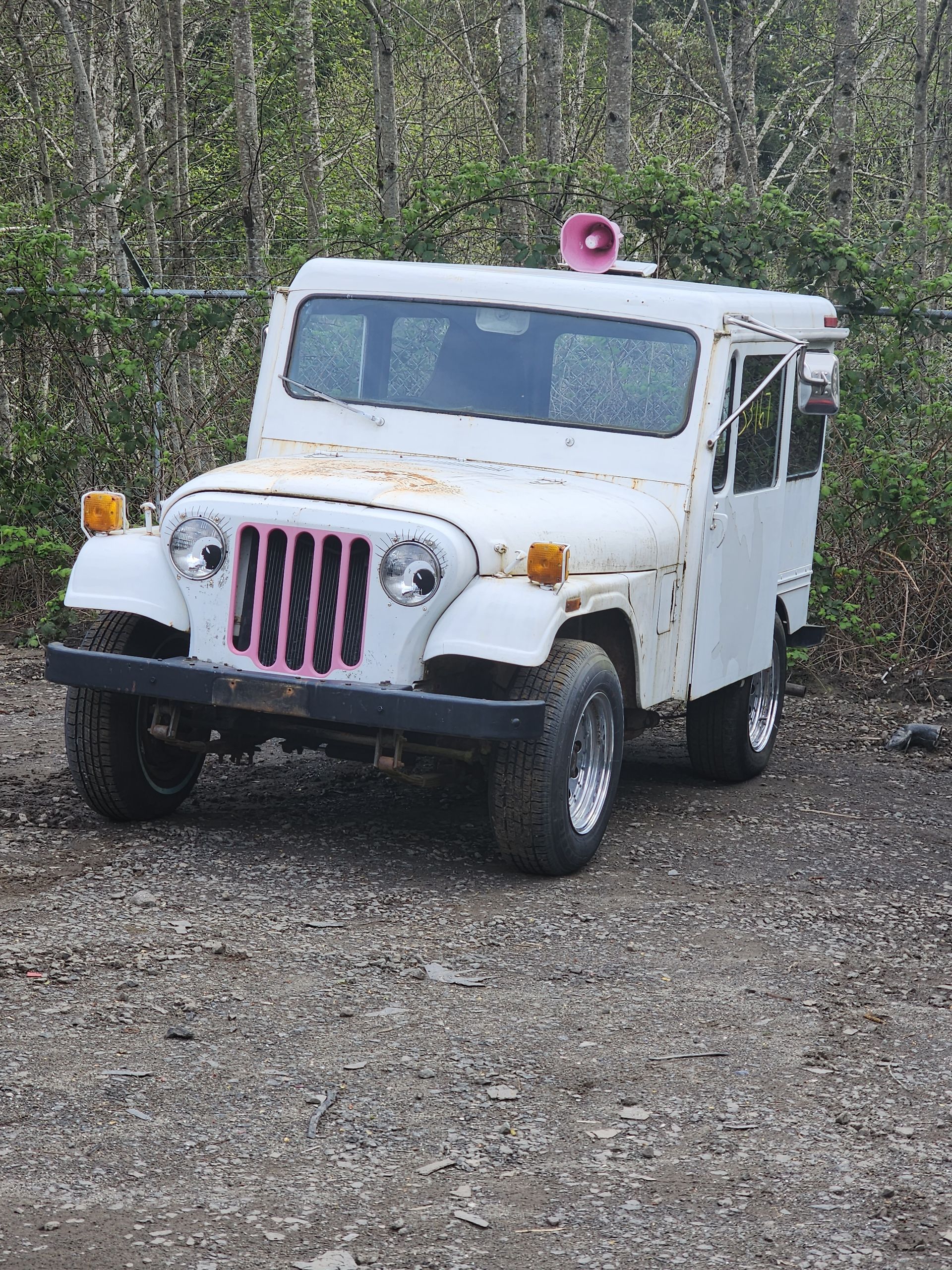 White ice cream truck on gravel, with a pink grille and siren on the roof.