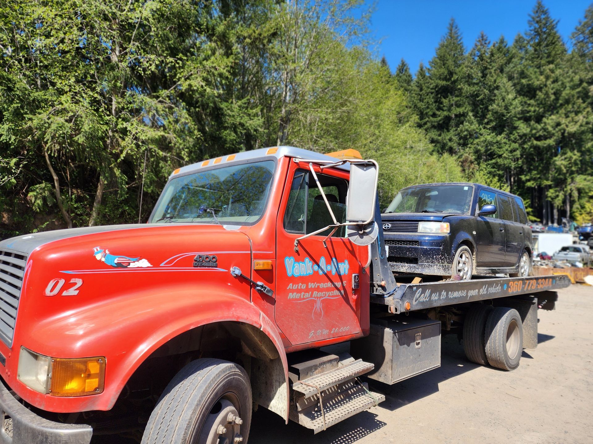 Red tow truck carrying a dark blue car on a sunny day in a wooded area.