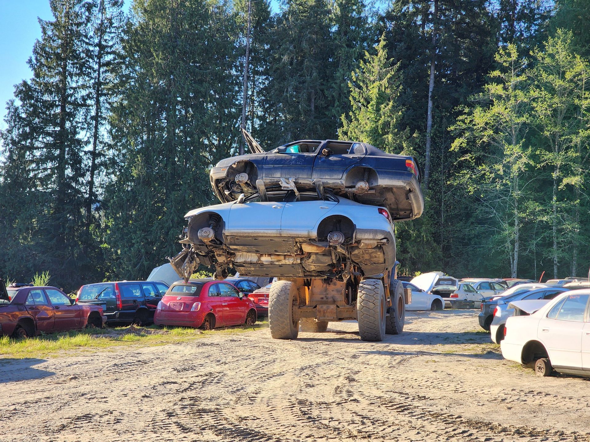 A large vehicle transports two salvaged cars, one on top of the other, in a junkyard.