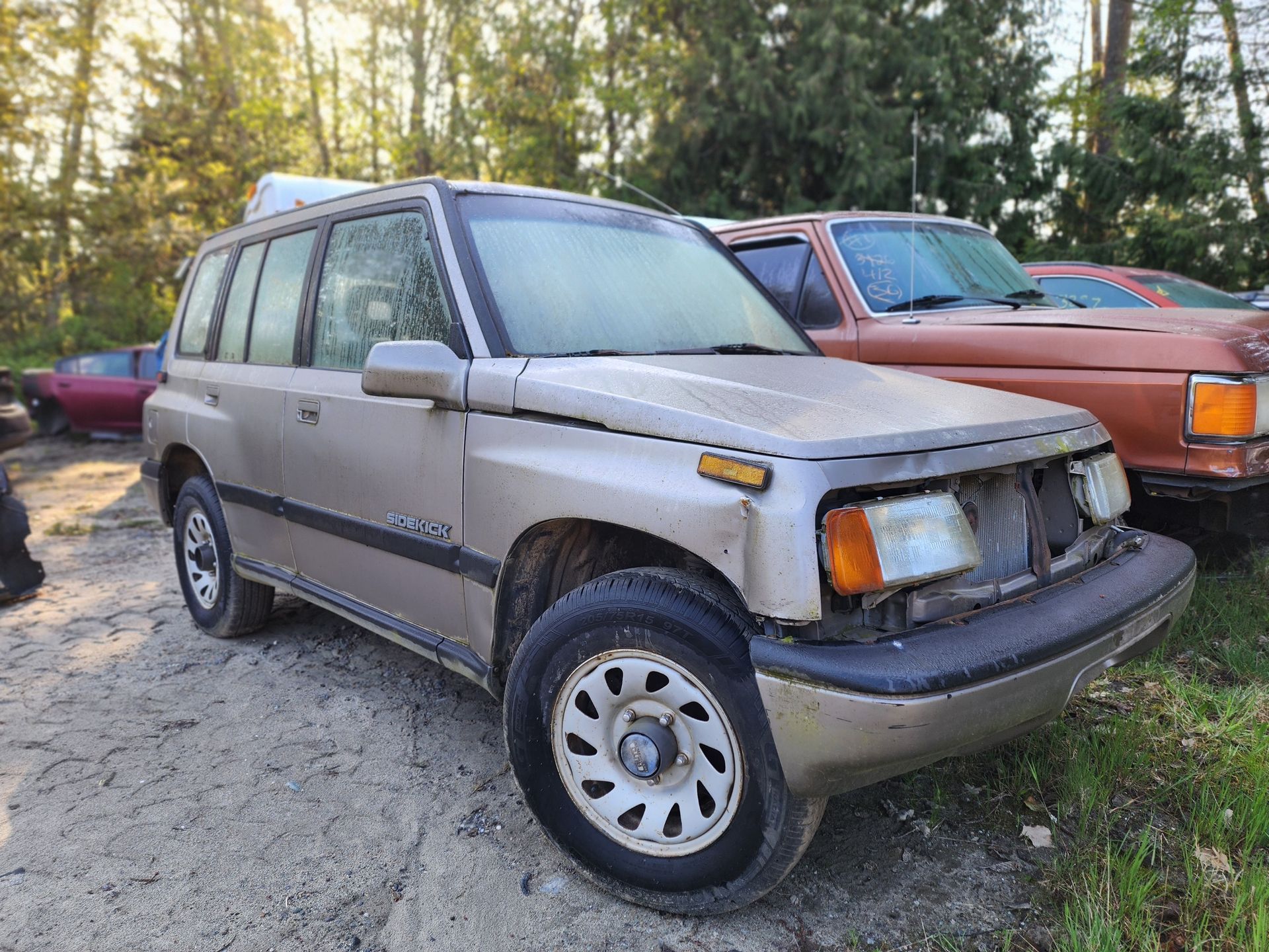 Tan Suzuki Sidekick SUV in a junkyard, parked next to a brown car, overcast day.