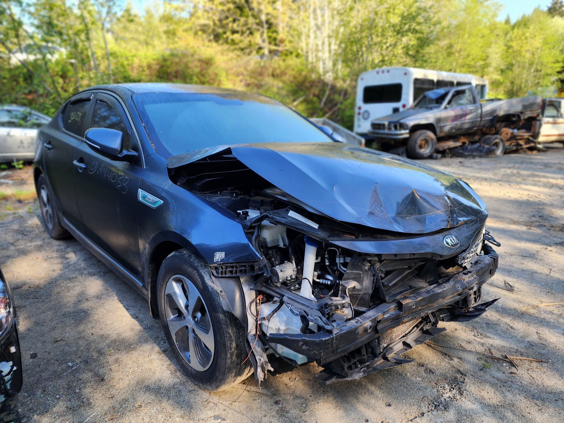 Damaged blue Kia Optima Hybrid car with front-end collision in a salvage yard.