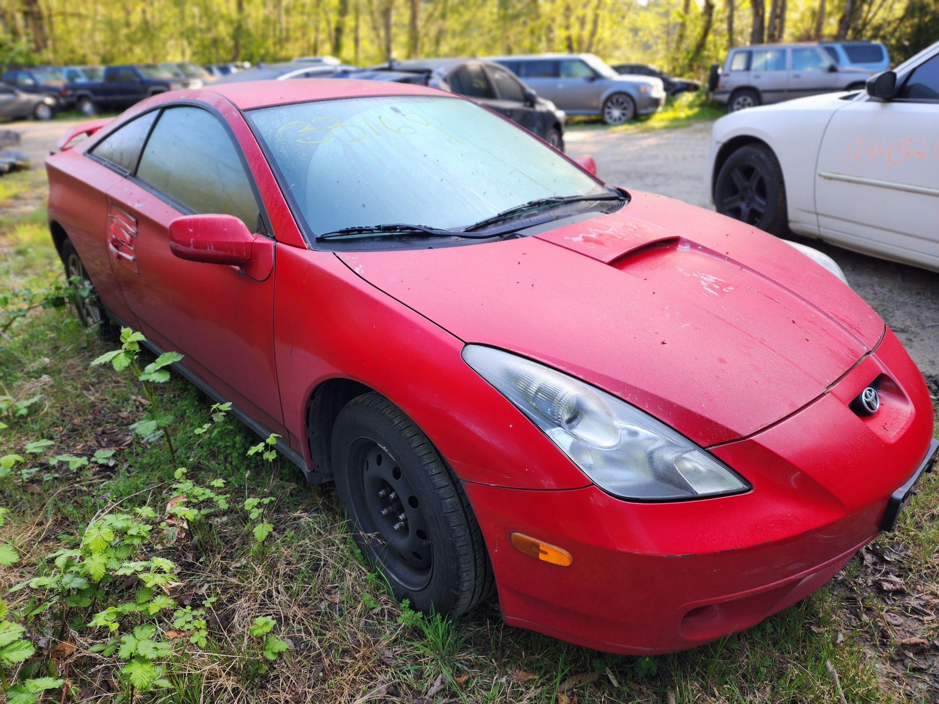 Red Toyota Celica coupe in a junkyard, surrounded by other vehicles and overgrown grass.