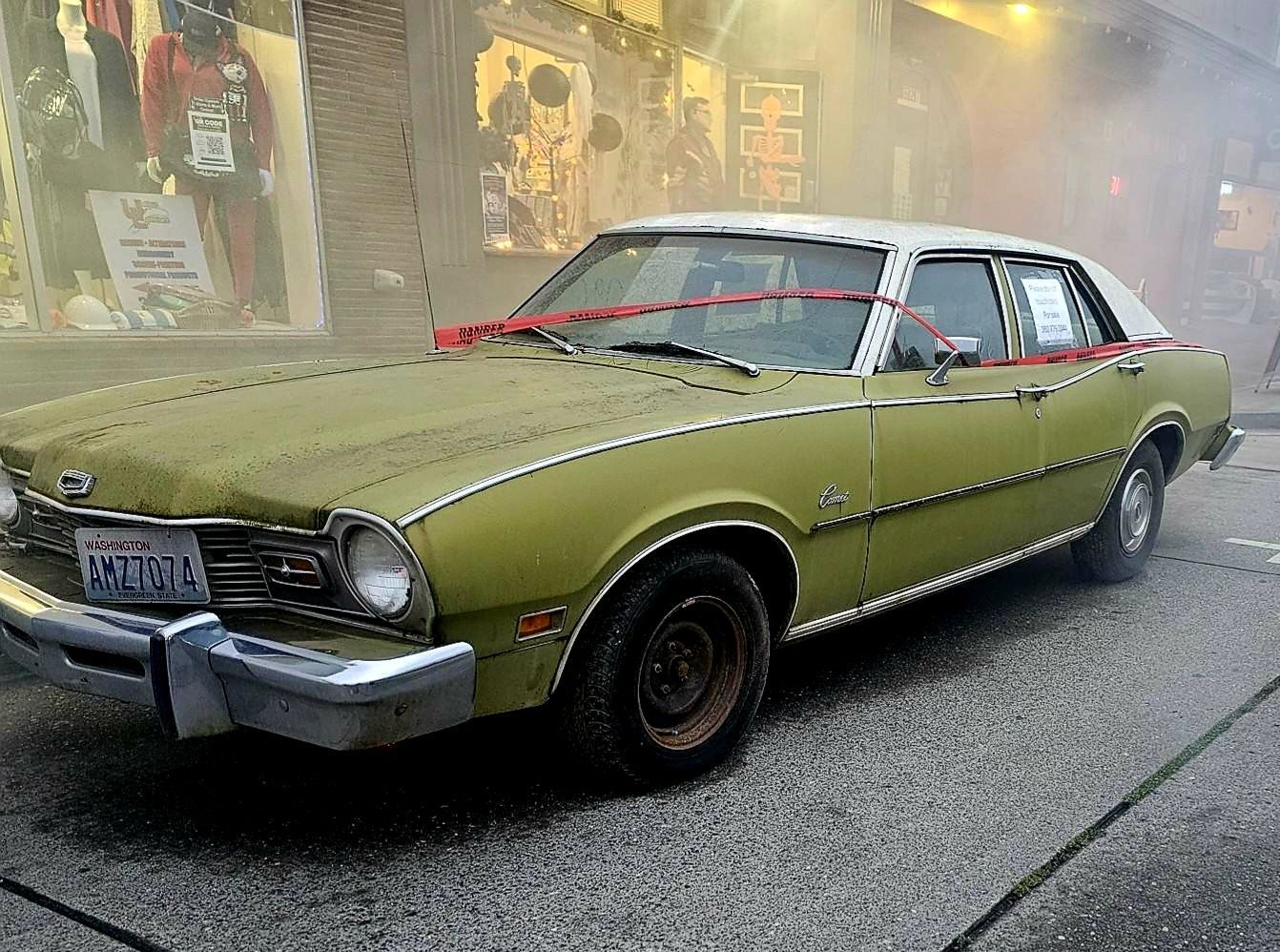 Green vintage car parked on a street with shop windows in the background.