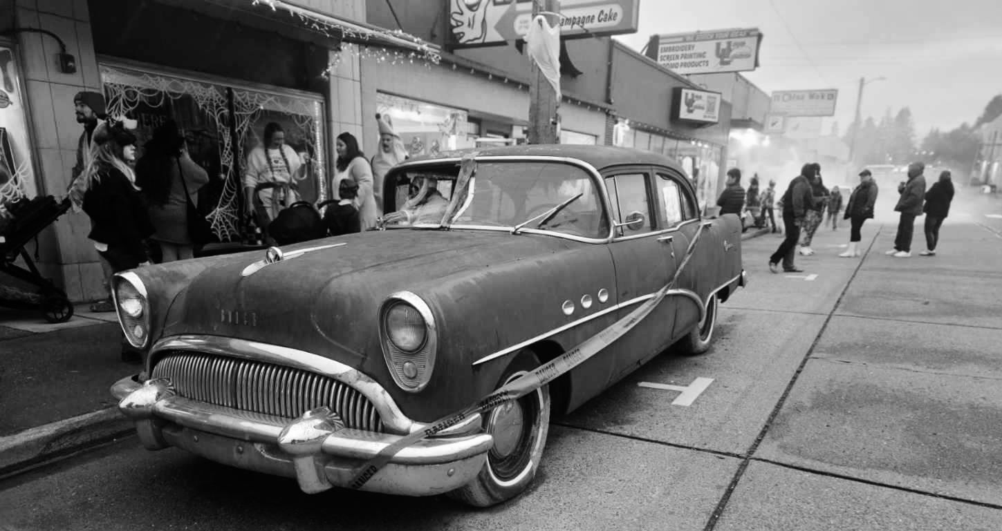 Black and white photo of a vintage Buick car parked on a street with people and shops in the background.