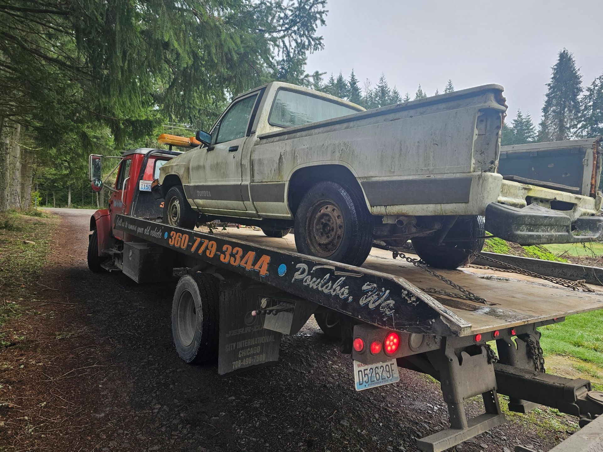 A weathered pickup truck loaded onto a red tow truck on a gravel road.