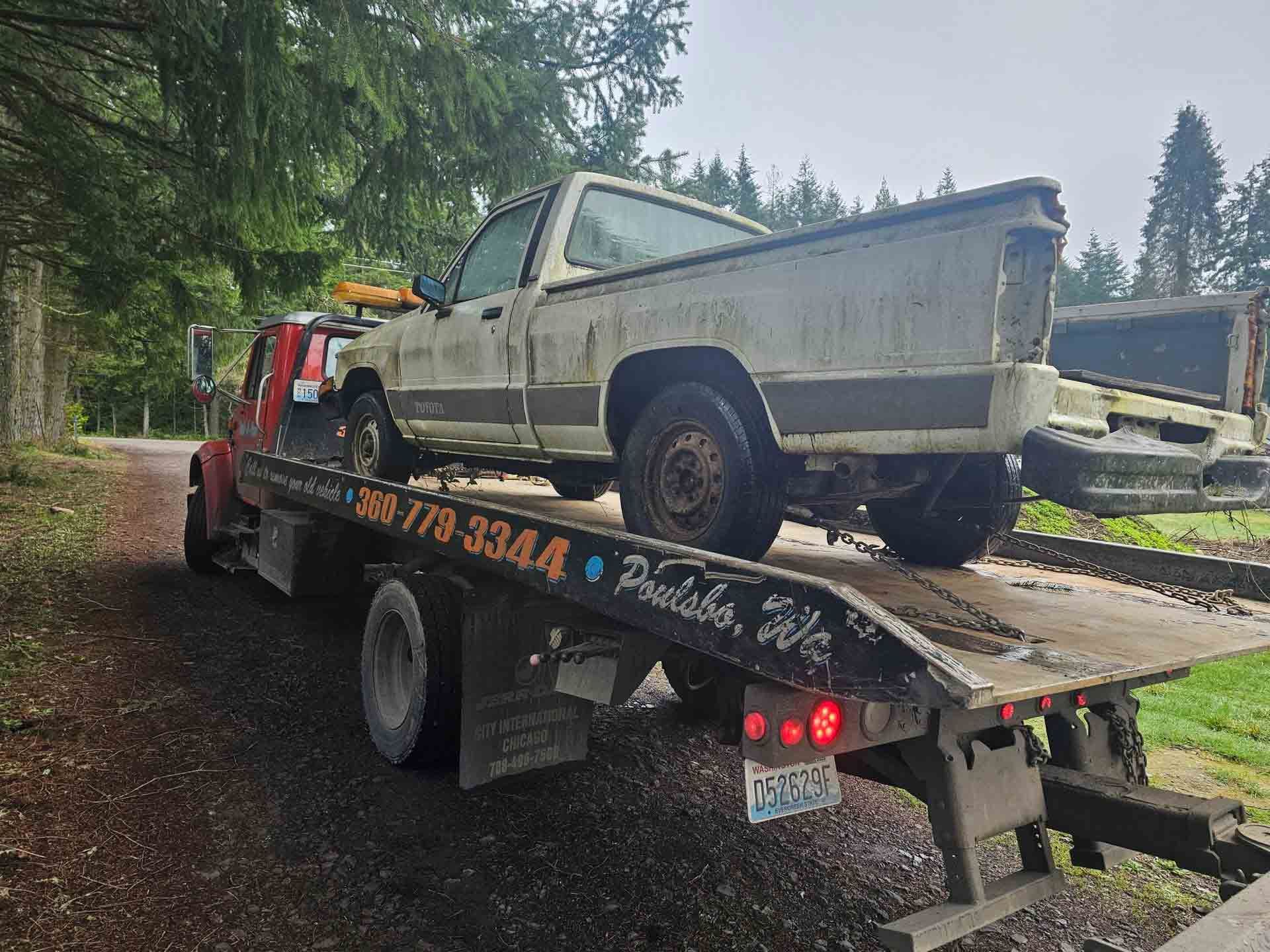 A weathered pickup truck loaded onto a red tow truck on a gravel road.