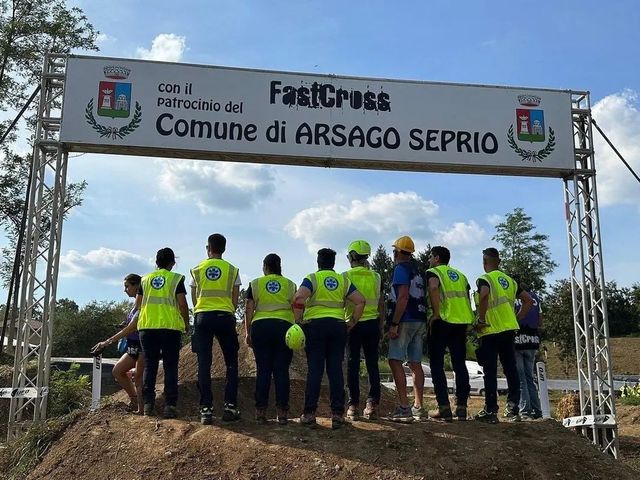 Gruppo di persone con giubbotti fluorescenti sotto uno striscione della gara in un'area di arrivo all'aperto, viste da dietro.