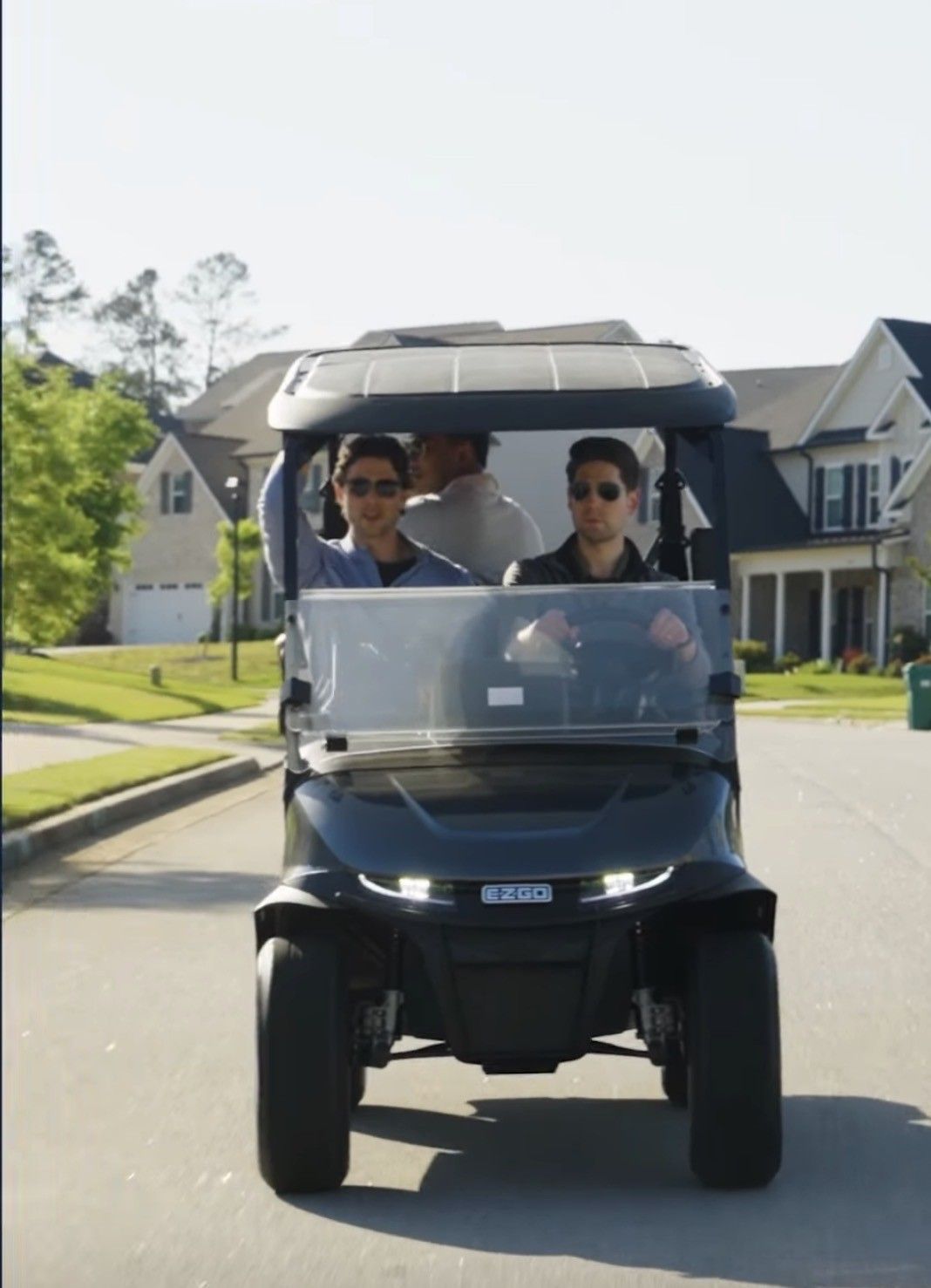Two people wearing sunglasses ride in a black golf cart on a residential street during the day.
