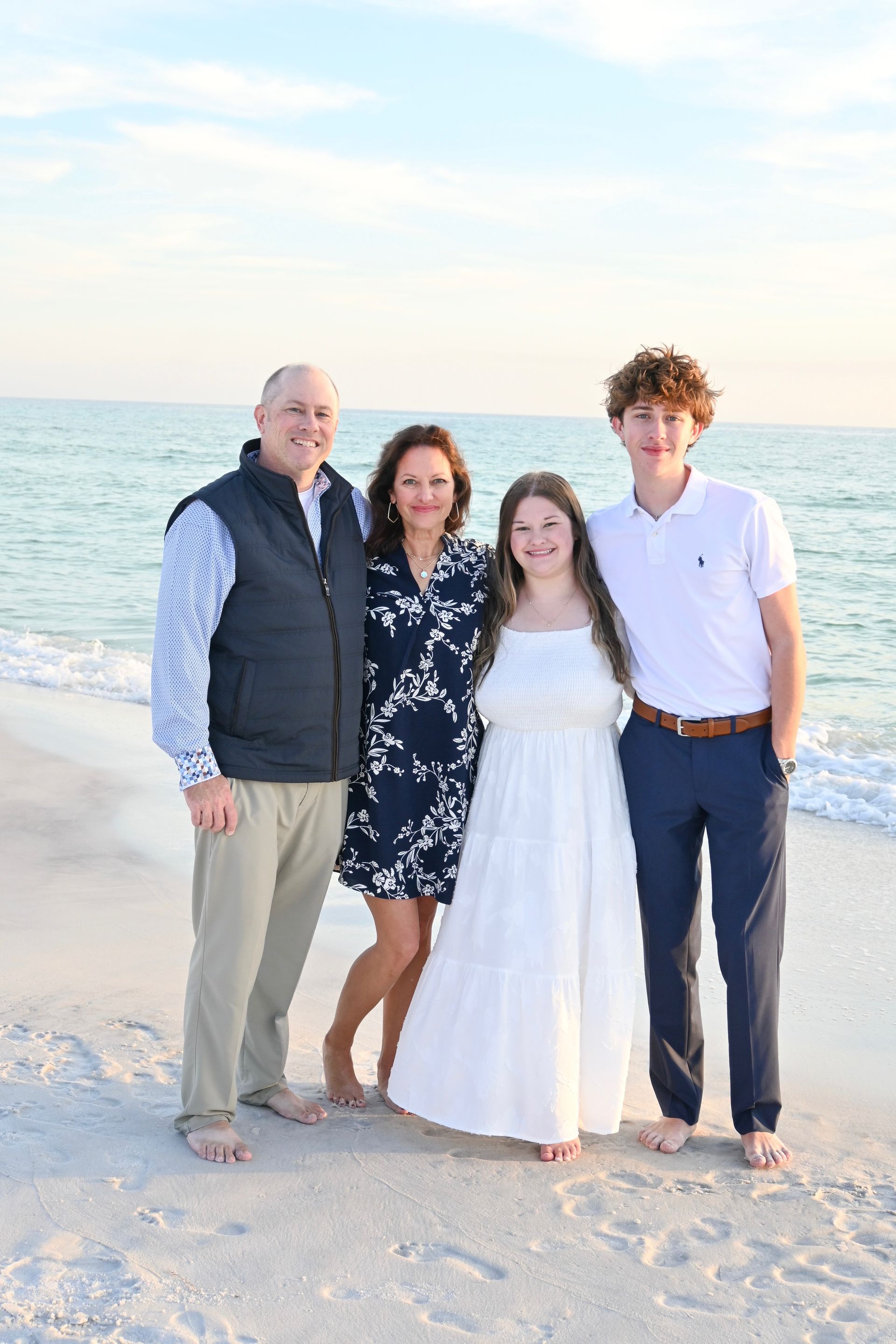A family of four stands together on a sandy beach at sunset, smiling toward the camera near the ocean water.
