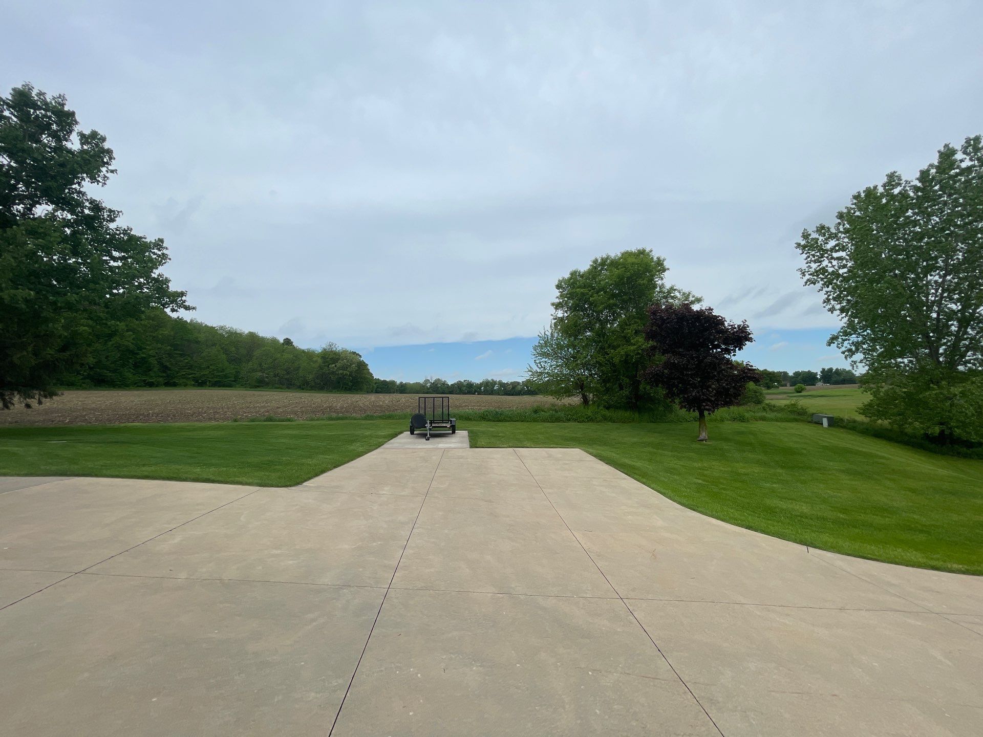 A concrete driveway leading to a grassy field with trees in the background.