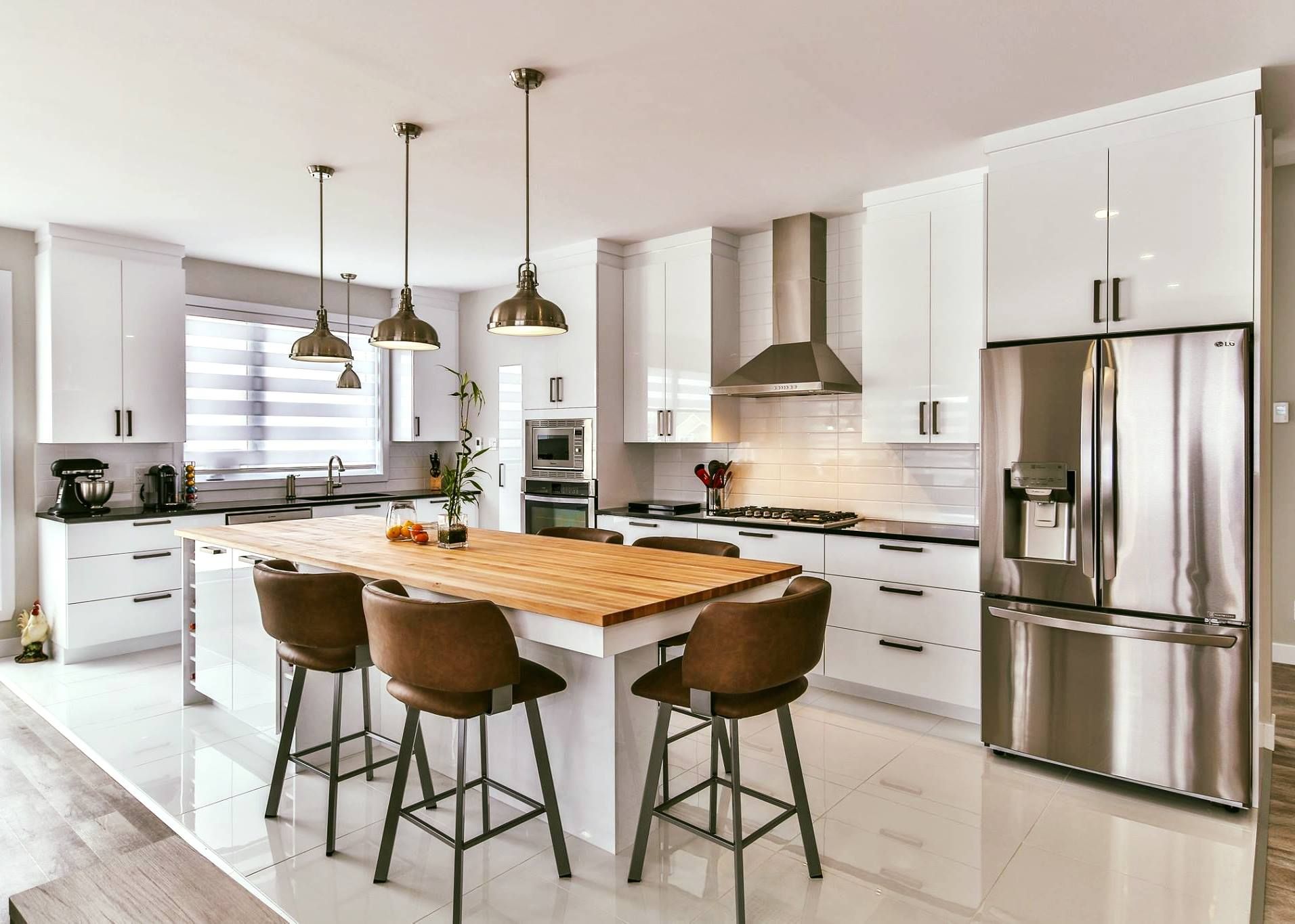 Cuisine blanche moderne avec îlot et tabourets marron, réfrigérateur en acier inoxydable et luminaires suspendus.