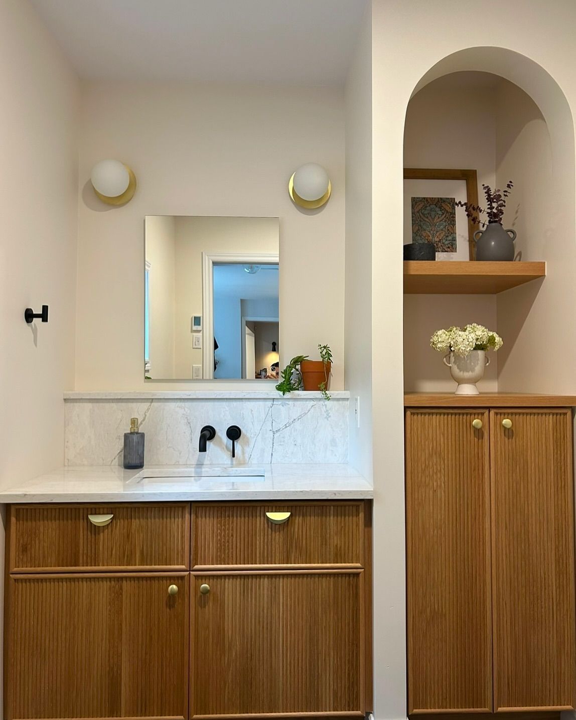 Salle de bain avec armoires en bois, comptoir en marbre, miroir et étagère arquée avec décor.