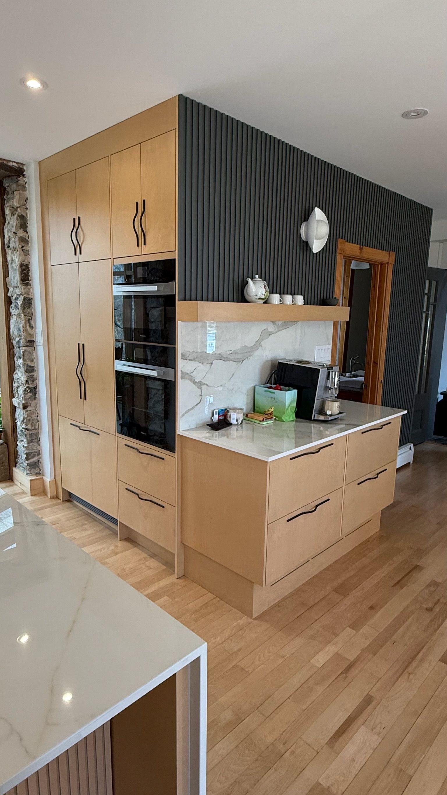 Cuisine moderne avec armoires en bois clair, fours noirs et mur gris foncé texturé.