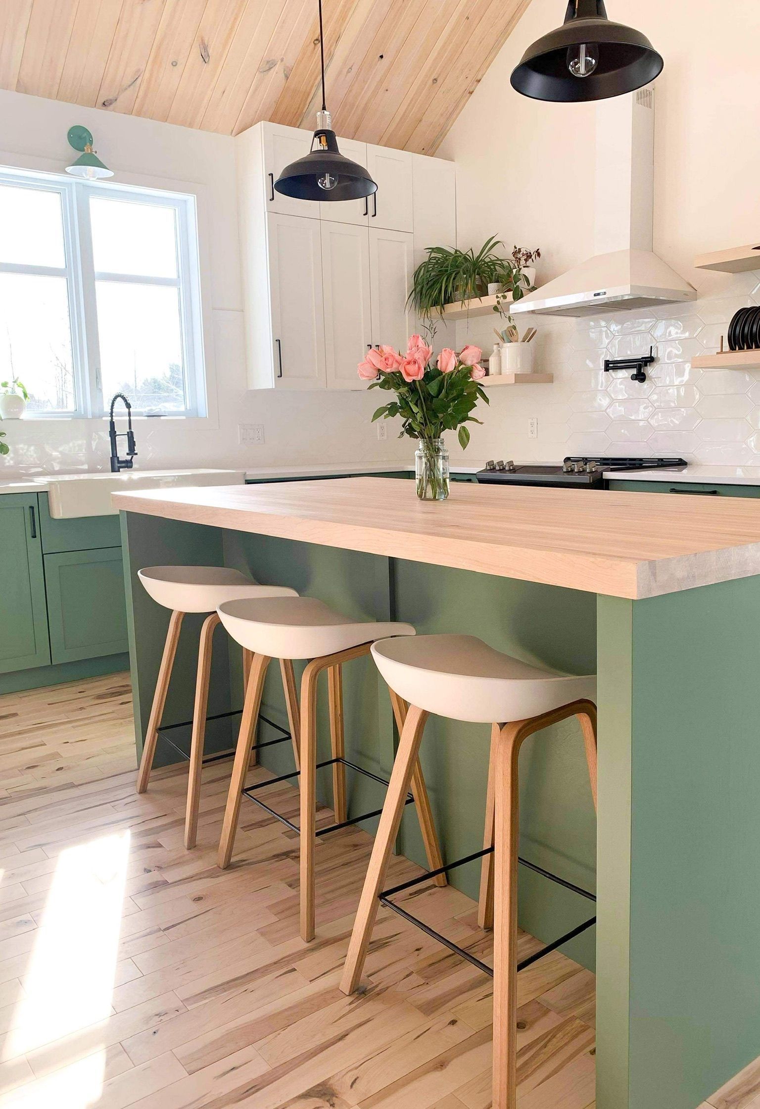 Cuisine verte avec îlot central, comptoirs en bois et tabourets. Fleurs roses, luminaires noirs et murs blancs.