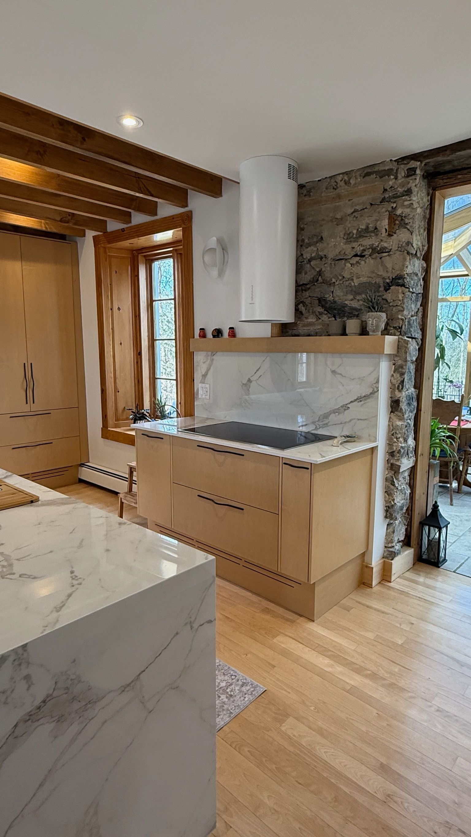 Cuisine avec parquet, mur en pierre et armoires en bois clair. Plan de travail en marbre et hotte blanche.