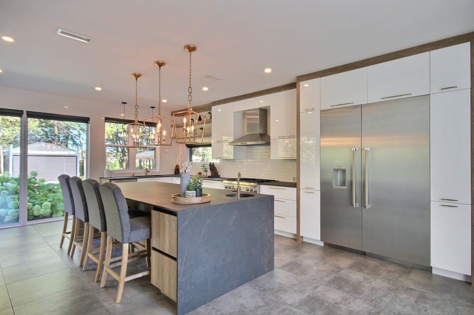 Cuisine moderne avec armoires blanches, îlot gris, réfrigérateur en acier inoxydable et luminaires suspendus dorés.