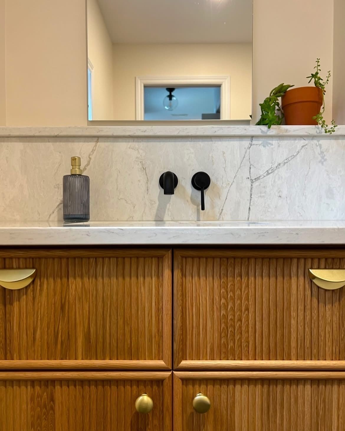 Meuble-lavabo avec armoires en bois, comptoir en marbre, luminaires noirs et une plante.