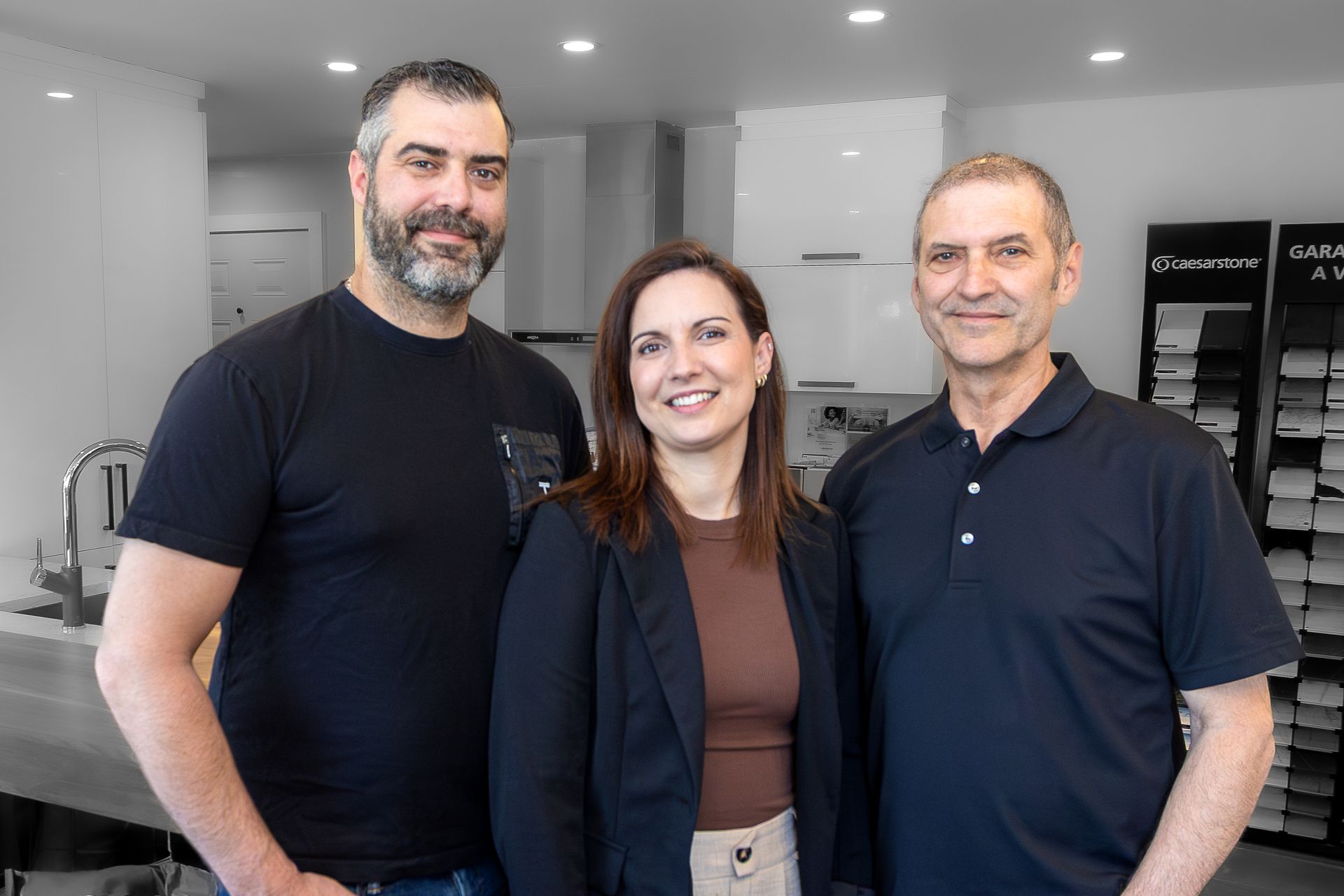 Trois personnes souriantes dans une salle d'exposition ; deux hommes encadrant une femme. Éclairage tamisé, fond blanc et gris moderne.