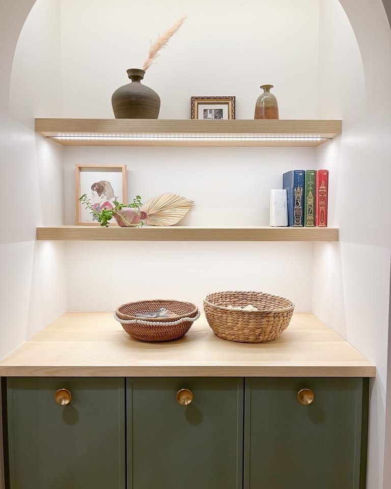 Étagères encastrées avec décoration : vases, livres, œuvres d'art encadrées et paniers. Armoires vertes avec boutons dorés.