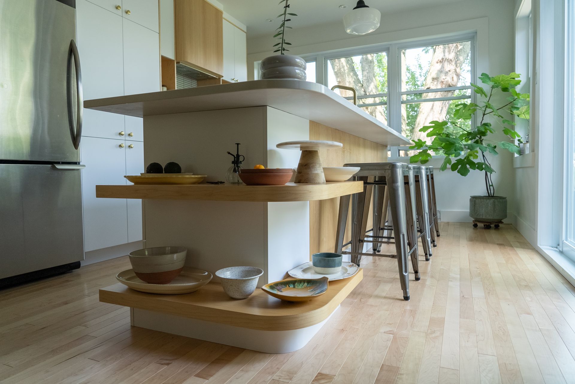 Îlot de cuisine moderne avec étagères en bois contenant des bols et des tabourets, à côté d'une fenêtre avec lumière naturelle.