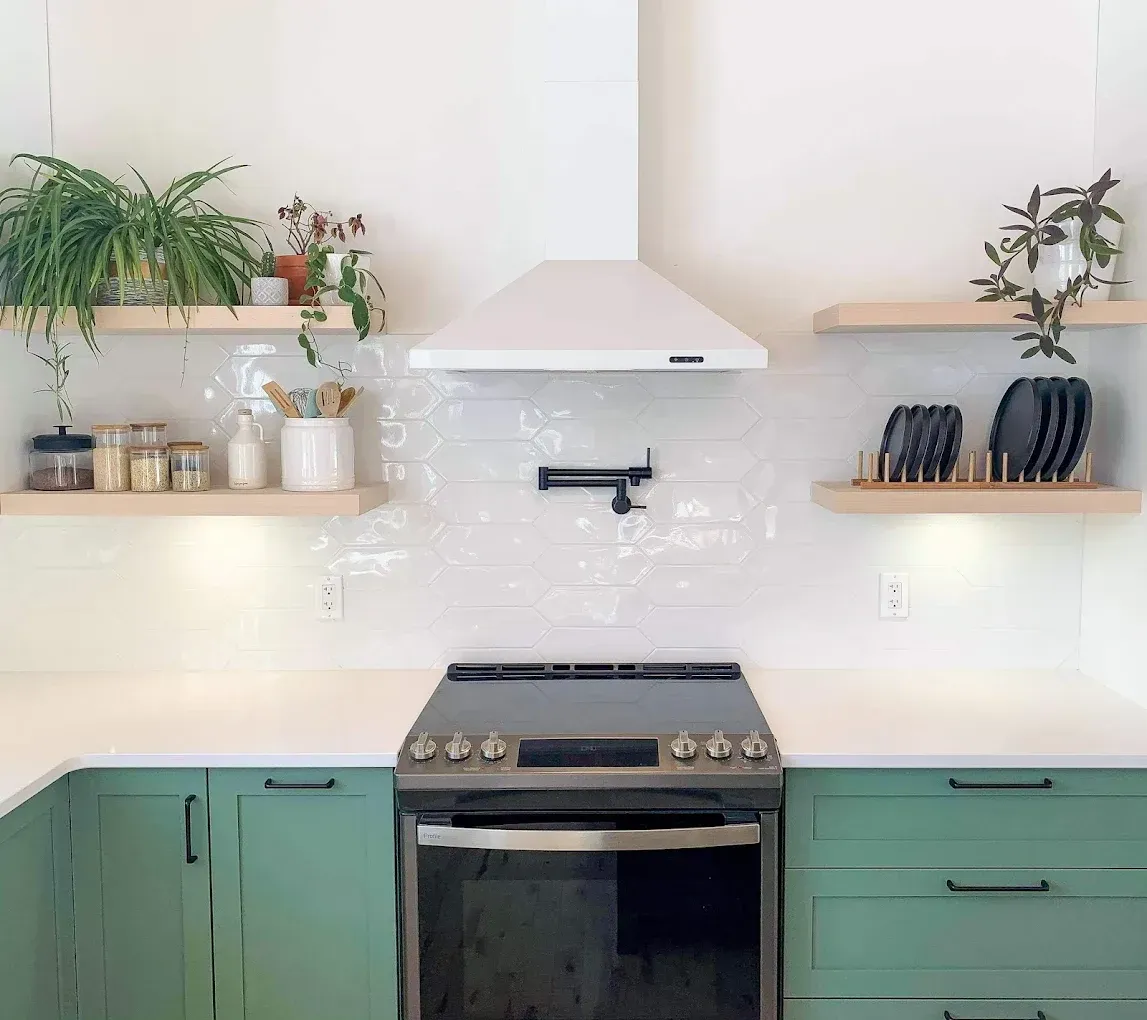 Cuisine verte avec cuisinière, hotte blanche, étagères ouvertes et plantes.