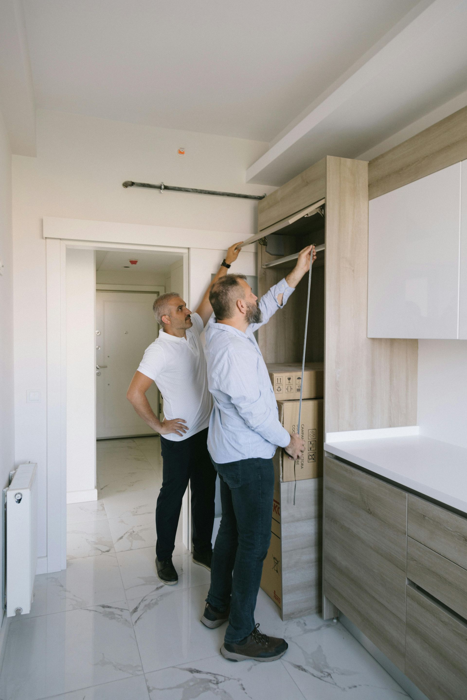 Deux hommes installent une armoire dans une pièce lumineuse avec une porte ouverte et des armoires blanches.