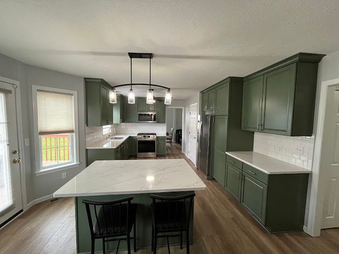 Green kitchen with white countertops, island with two black chairs, and modern lighting.