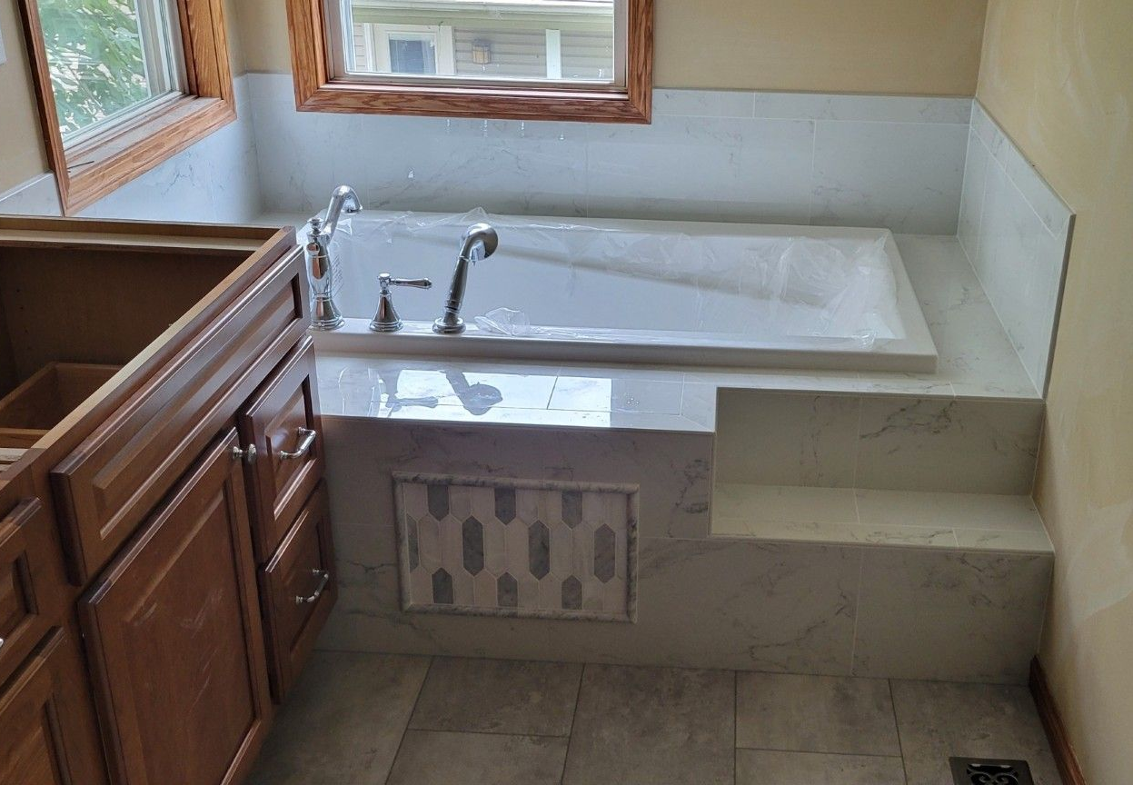 Bathroom with built-in bathtub, light marble tile, wooden vanity, and window.