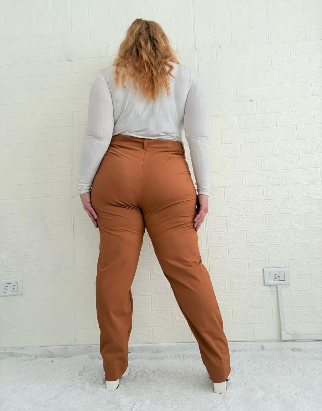 Mujer con top blanco y pantalón negro posa junto a una pared de ladrillos blancos.